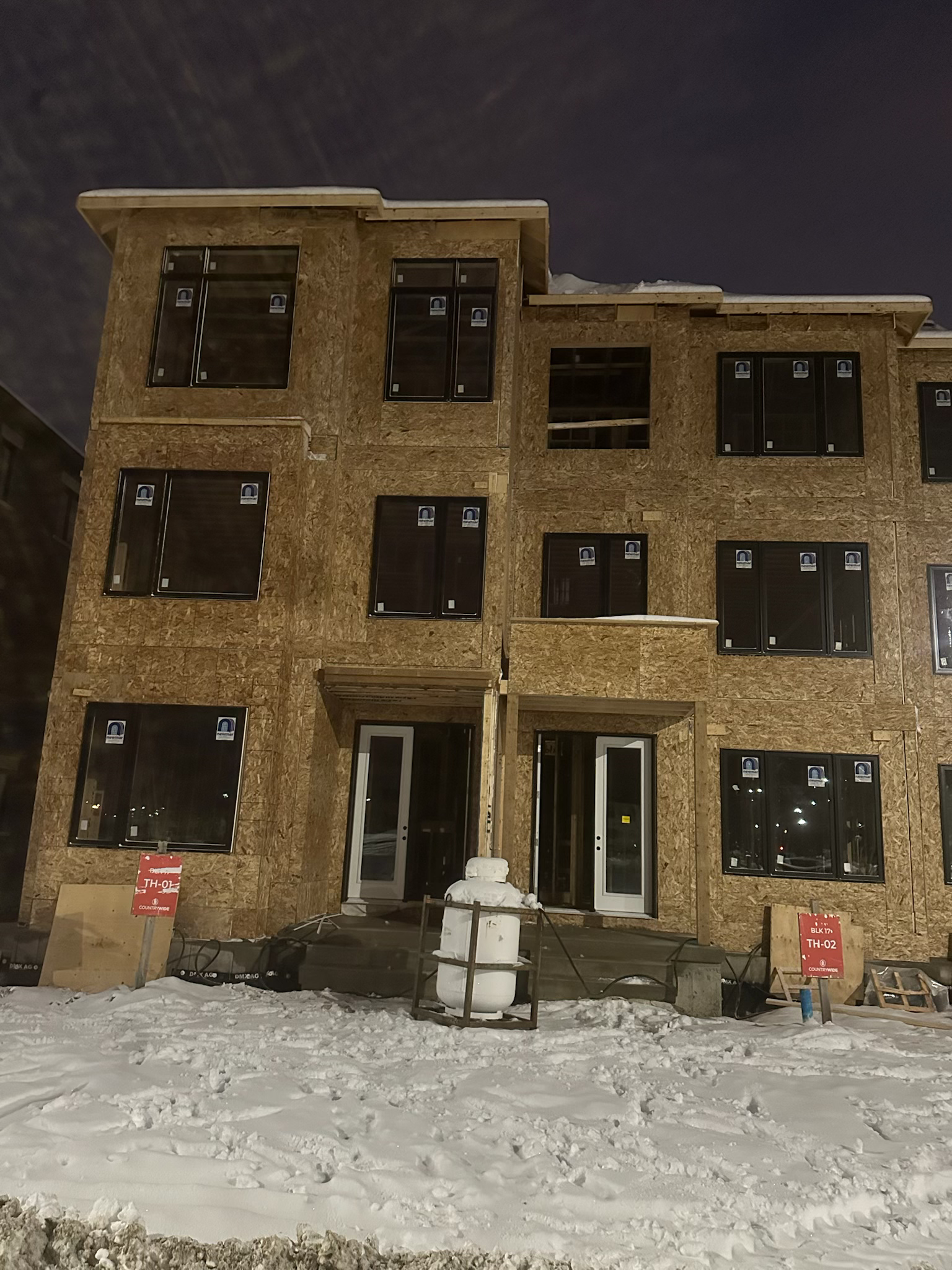 A multi-story building under construction with wood framing, large window openings, and snow on the ground at night.