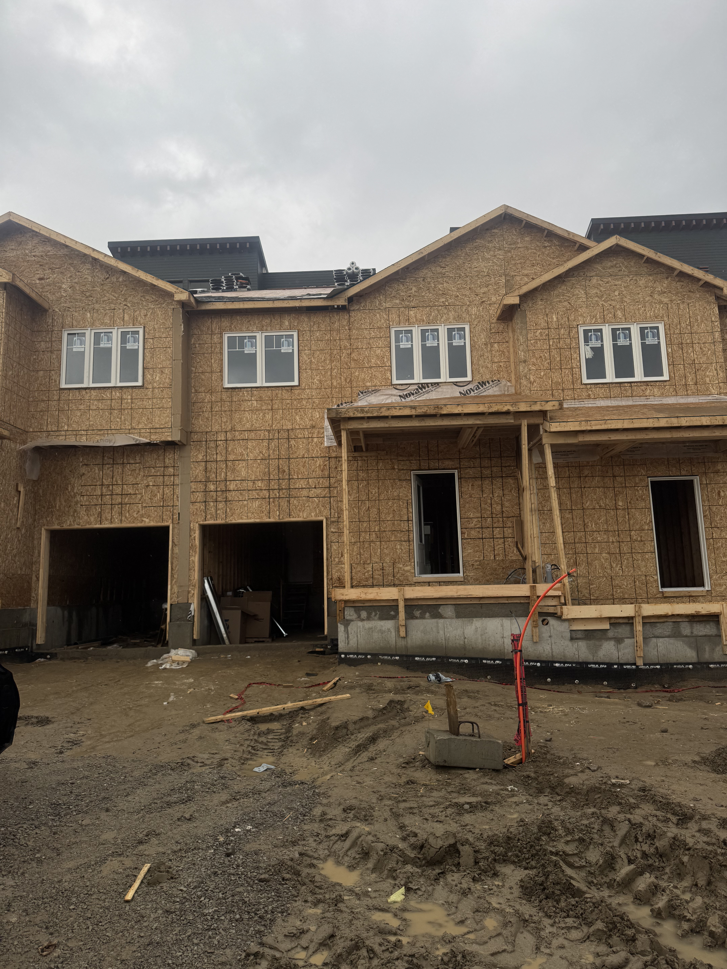 Front view of a house under construction with partially finished walls and roof, construction materials and equipment visible, muddy ground with tire tracks and construction debris.