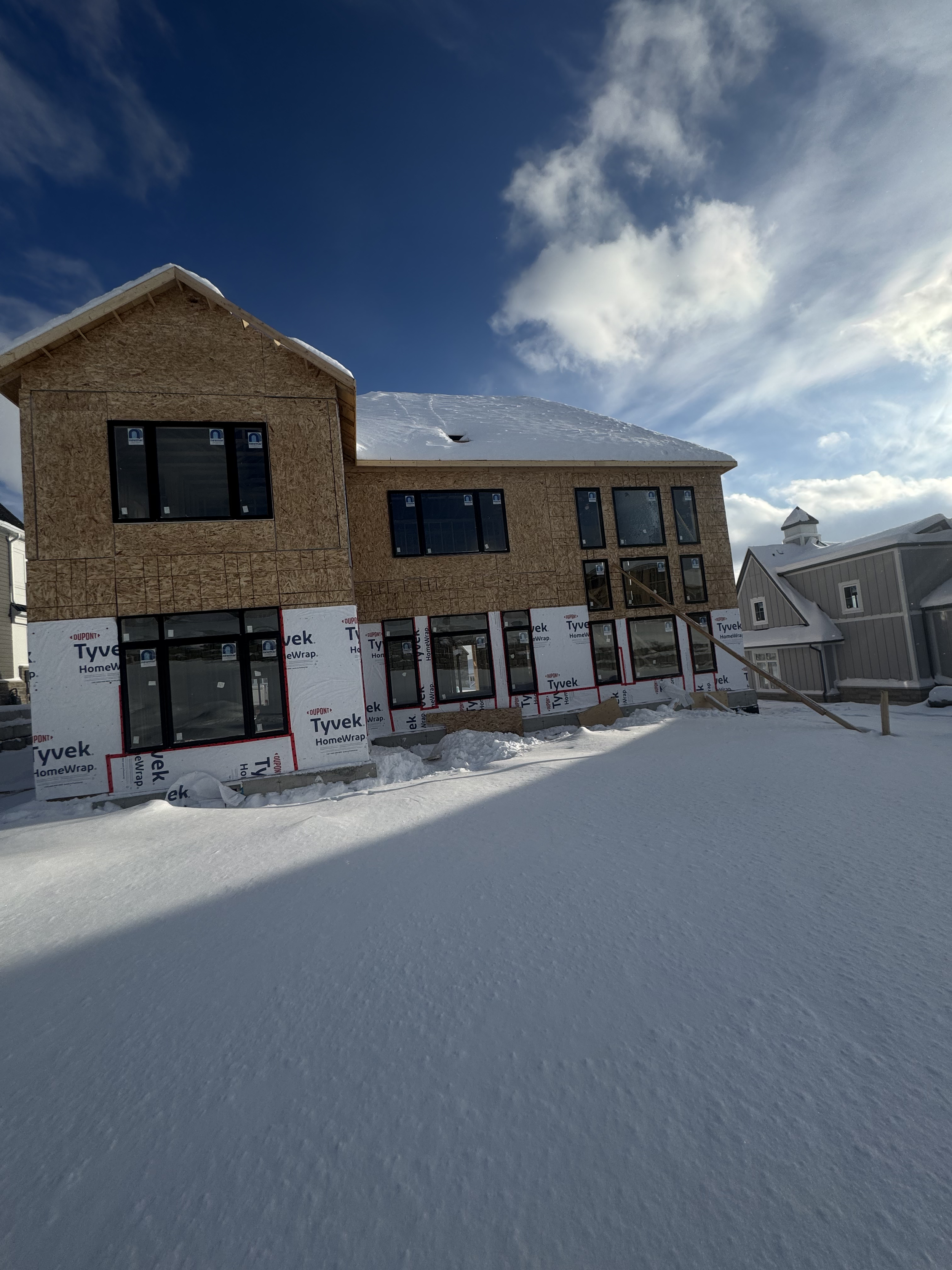 A two-story house under construction with exposed plywood and window frames, covered in snow, with a bright blue sky and scattered clouds above.