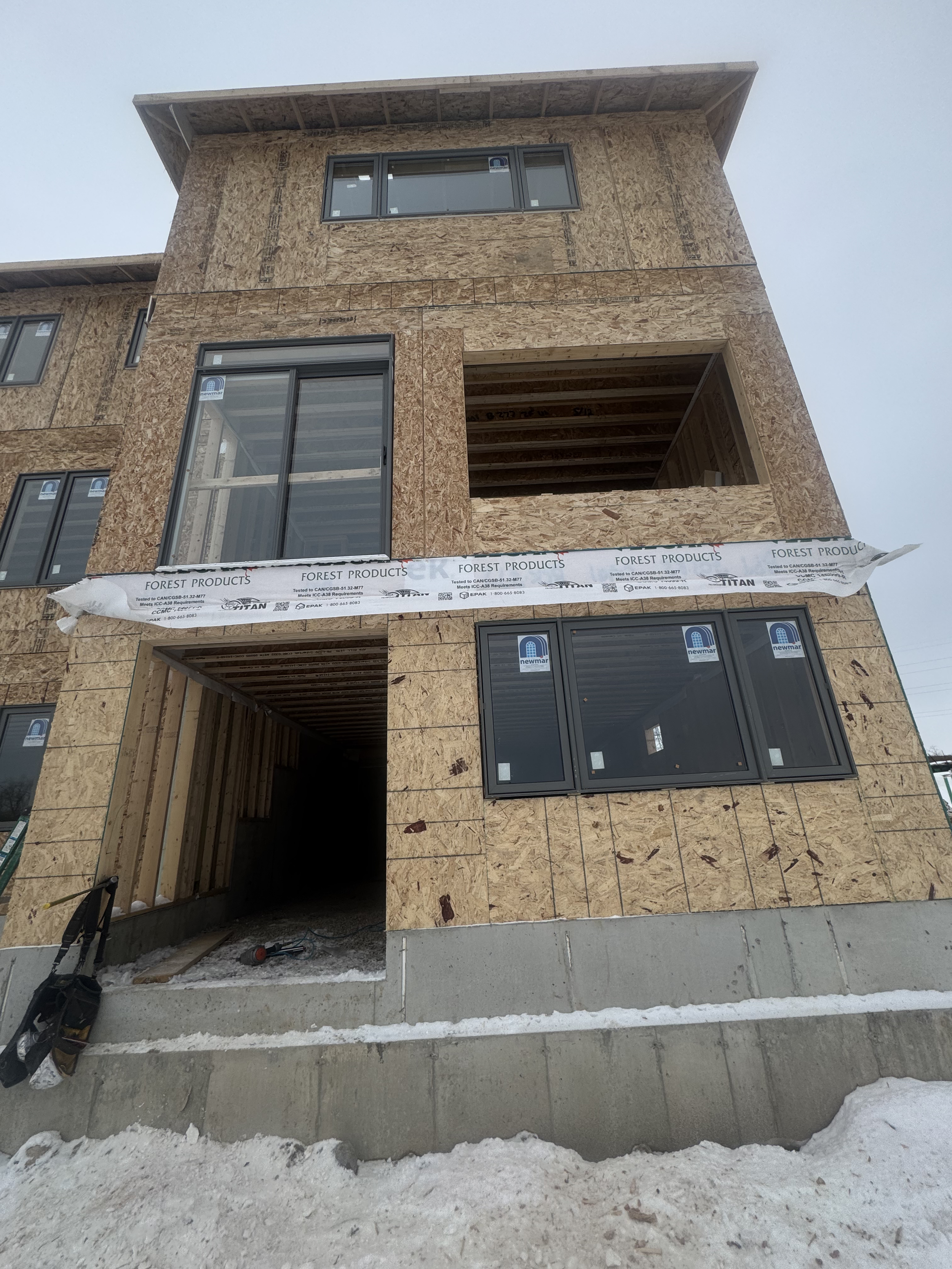 A multi-story building under construction with plywood sheathing and installed windows, partially covered with a construction barrier, with snow on the ground.