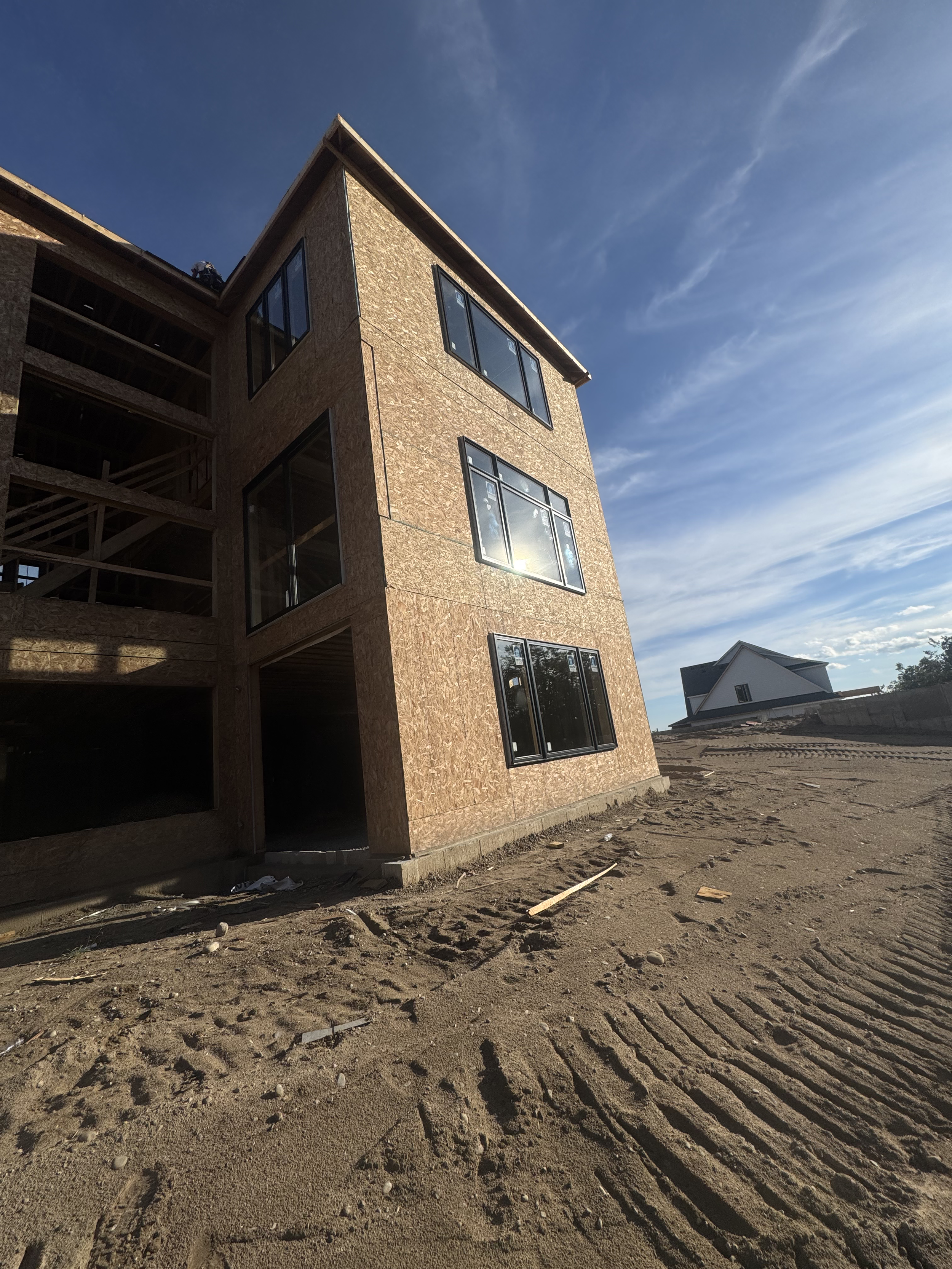 A multi-story house under construction with wood framing and large windows, set on dirt ground under a blue sky with clouds.