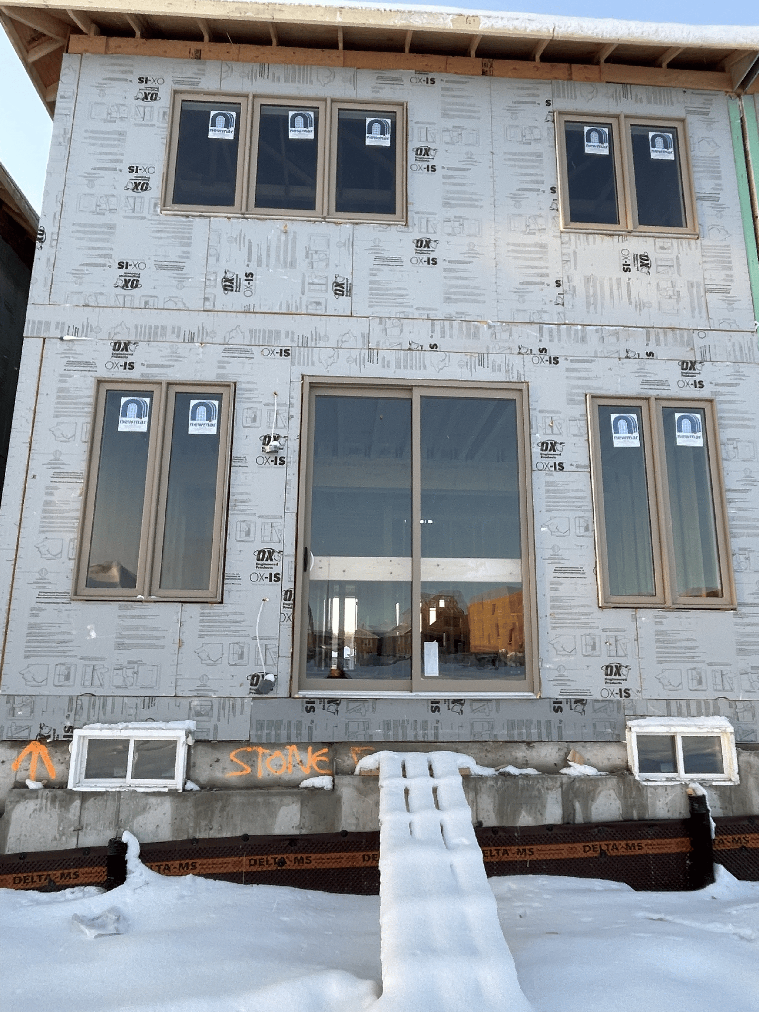 Front of a house under construction with snow on the ground, showing unfinished exterior with insulation panels, multiple windows, and a sliding glass door, with a small snow-covered ramp leading to the door.