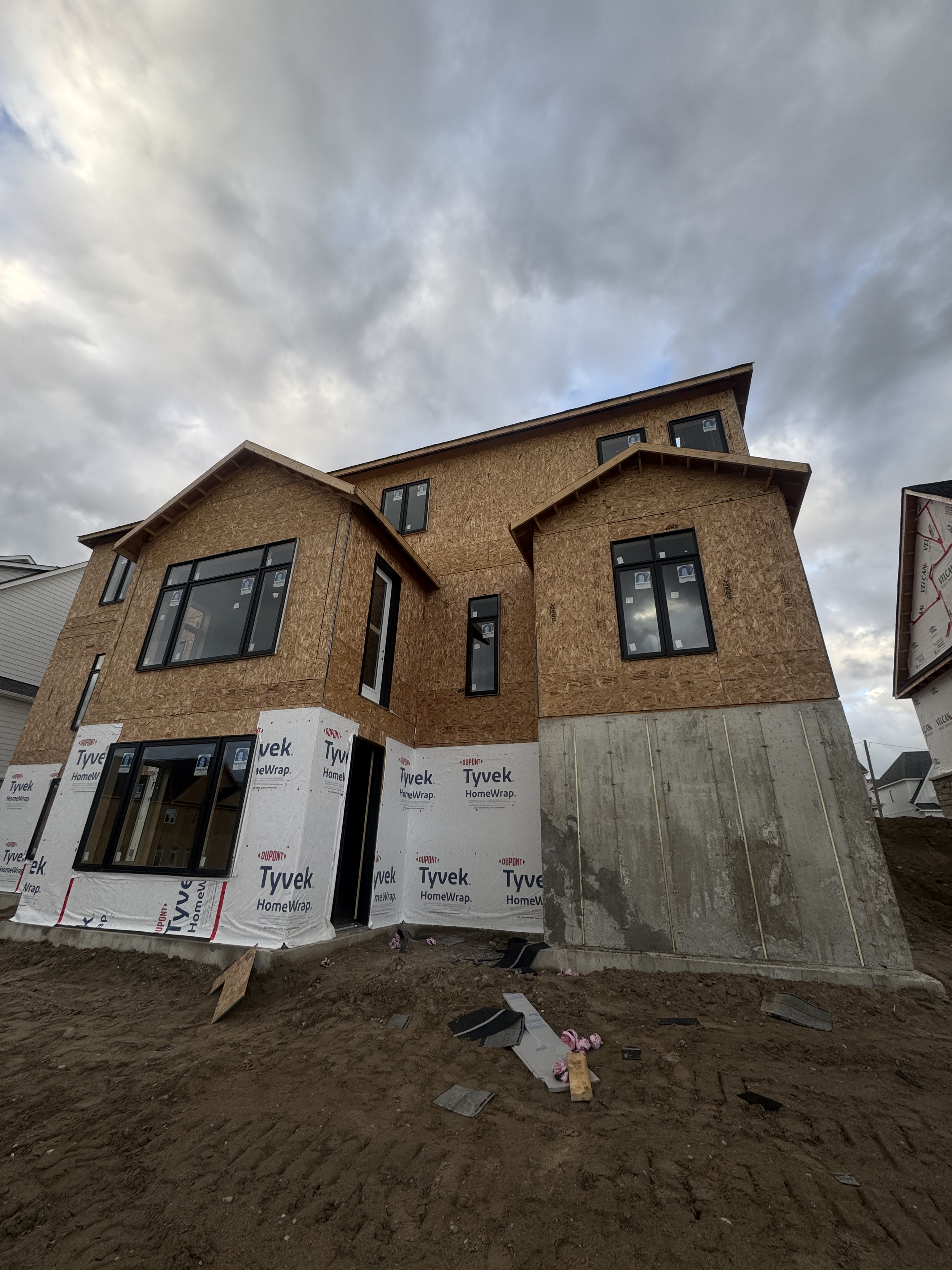 A multi-story house under construction with partially finished wooden exterior, large window openings, and a concrete foundation against a cloudy sky.