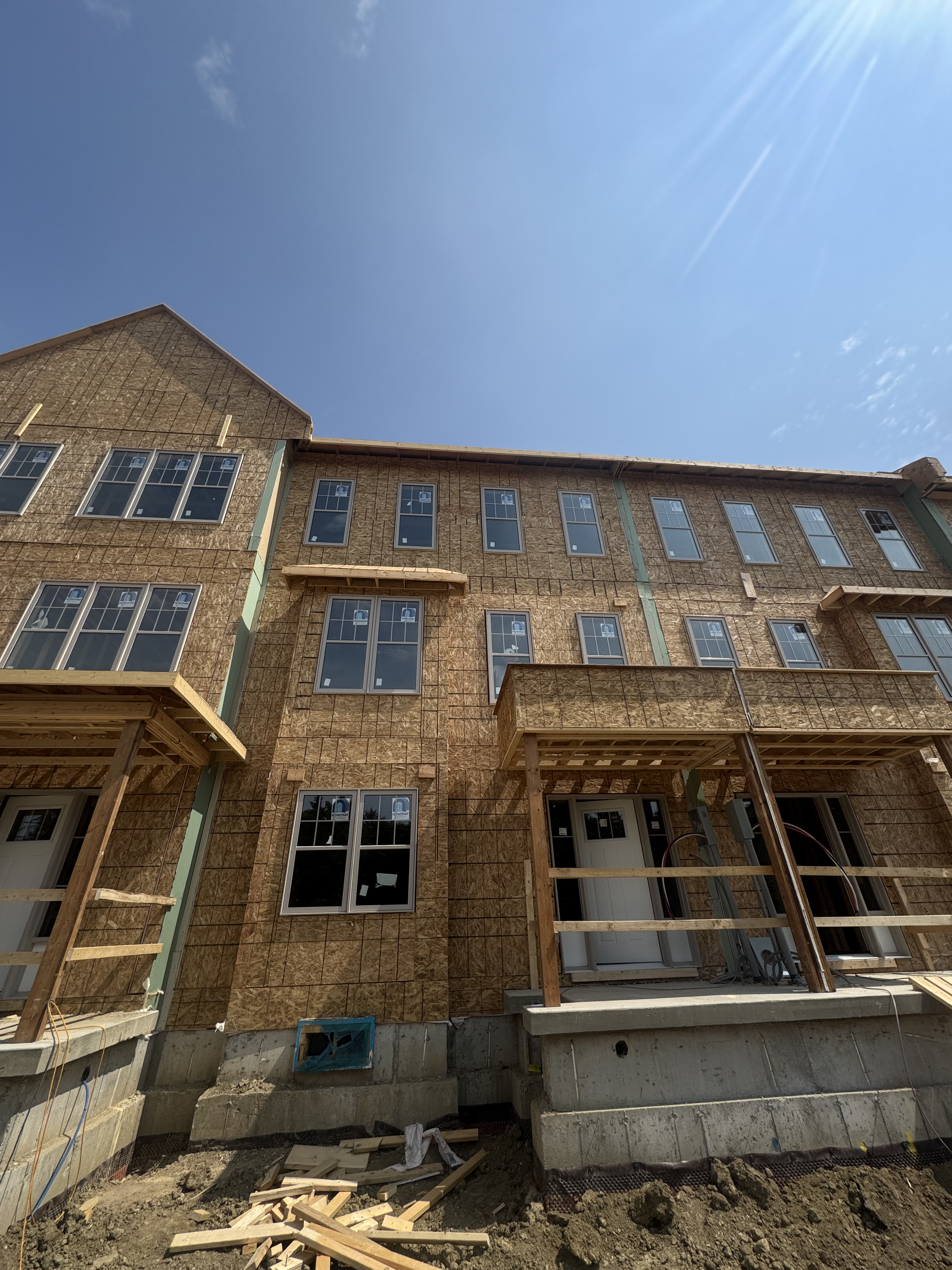 Under construction multi-story residential building with exposed plywood exterior, multiple windows, and wooden balconies, set against a clear blue sky.