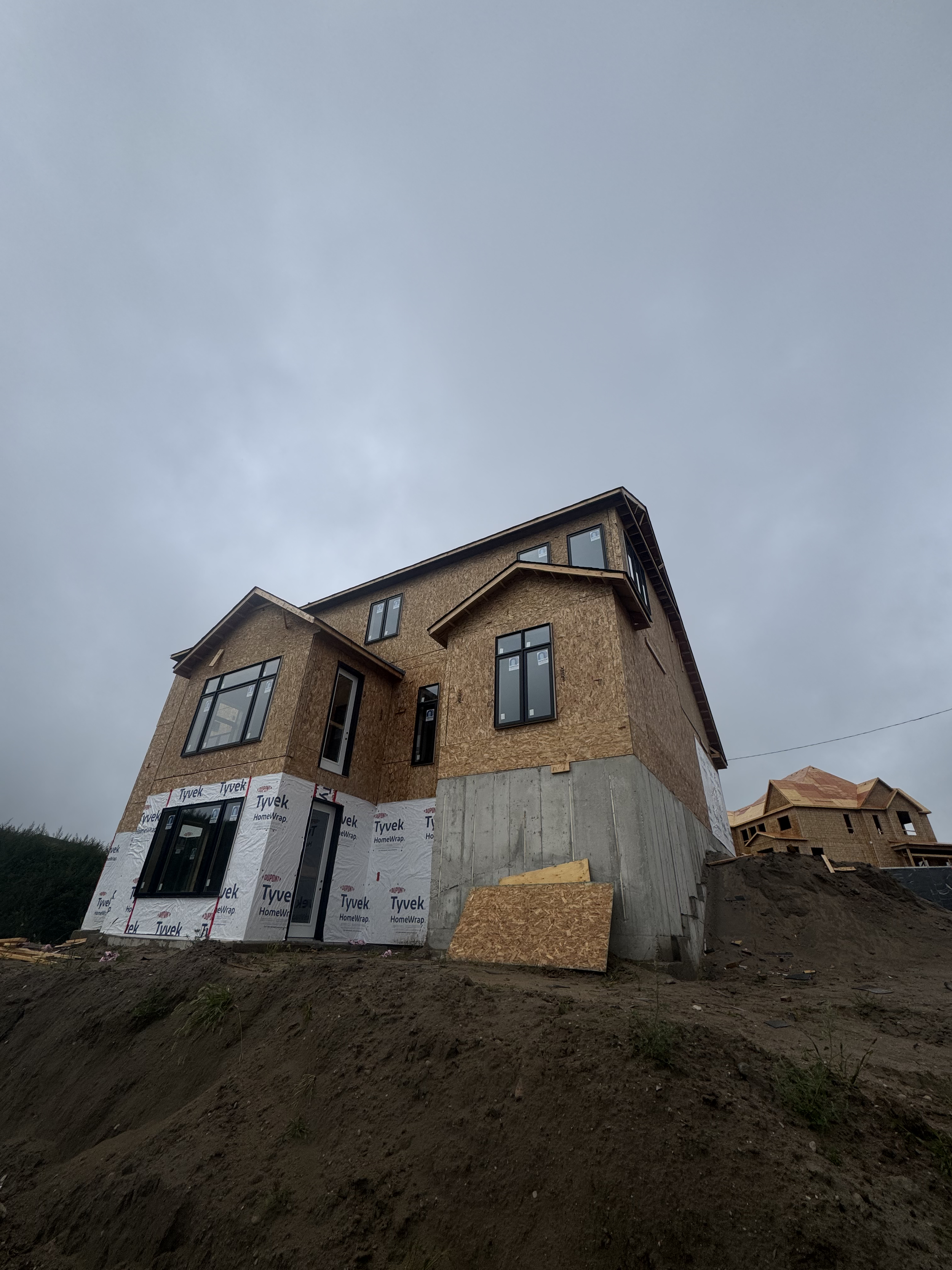 A partially constructed house on a sloped dirt lot under a cloudy sky.