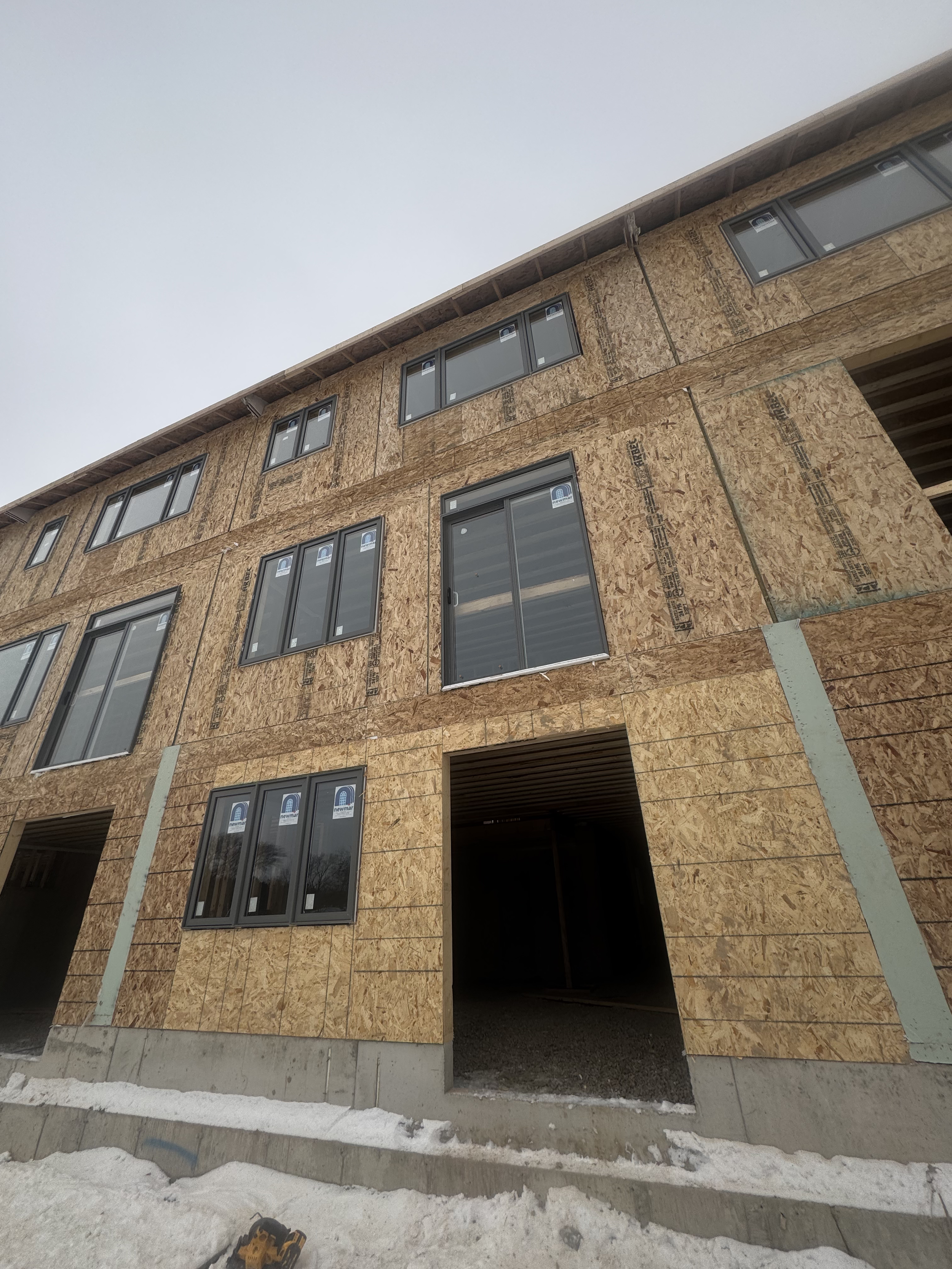Building under construction with plywood sheathing and multiple window openings, snow on the ground, and a cloudy sky.