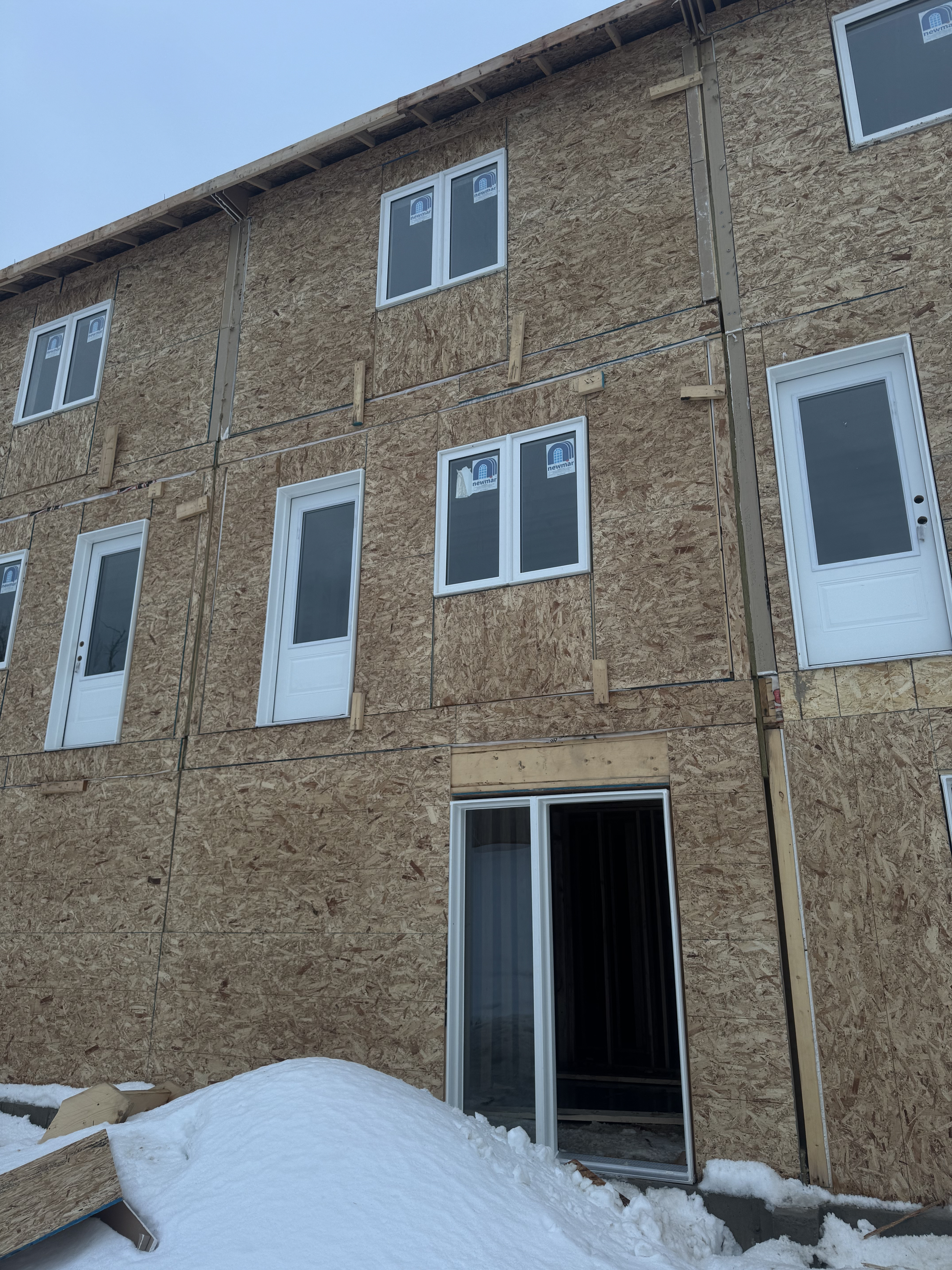Building under construction with OSB sheathing and white framed windows, snow on ground, clear sky.