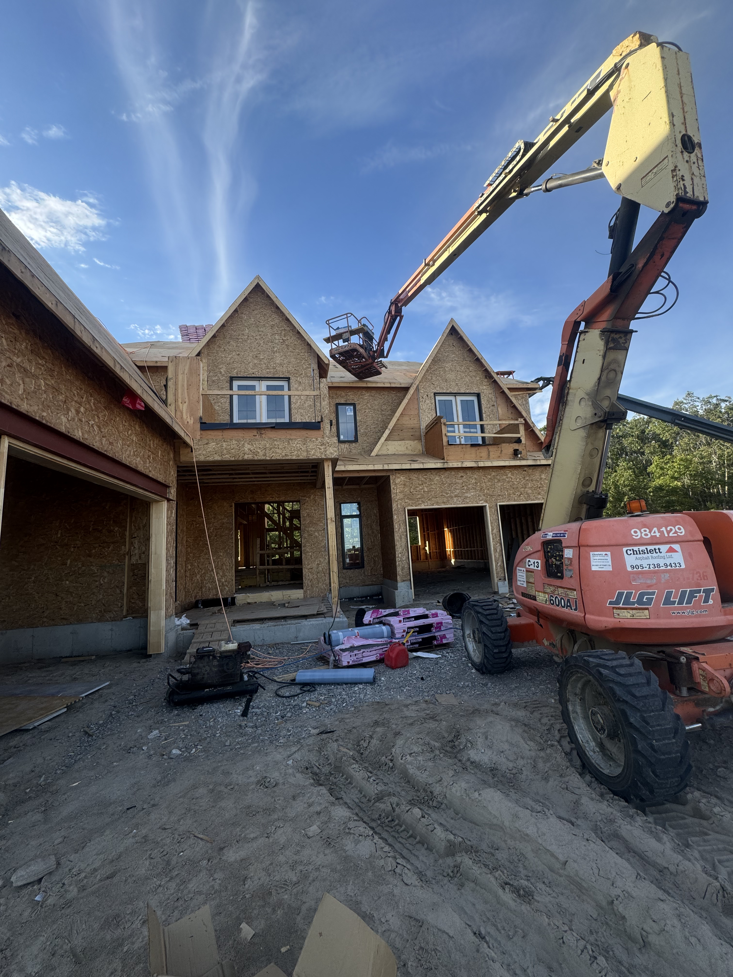 Construction site with a partially built two-story house, construction equipment, and materials, under a blue sky with clouds.