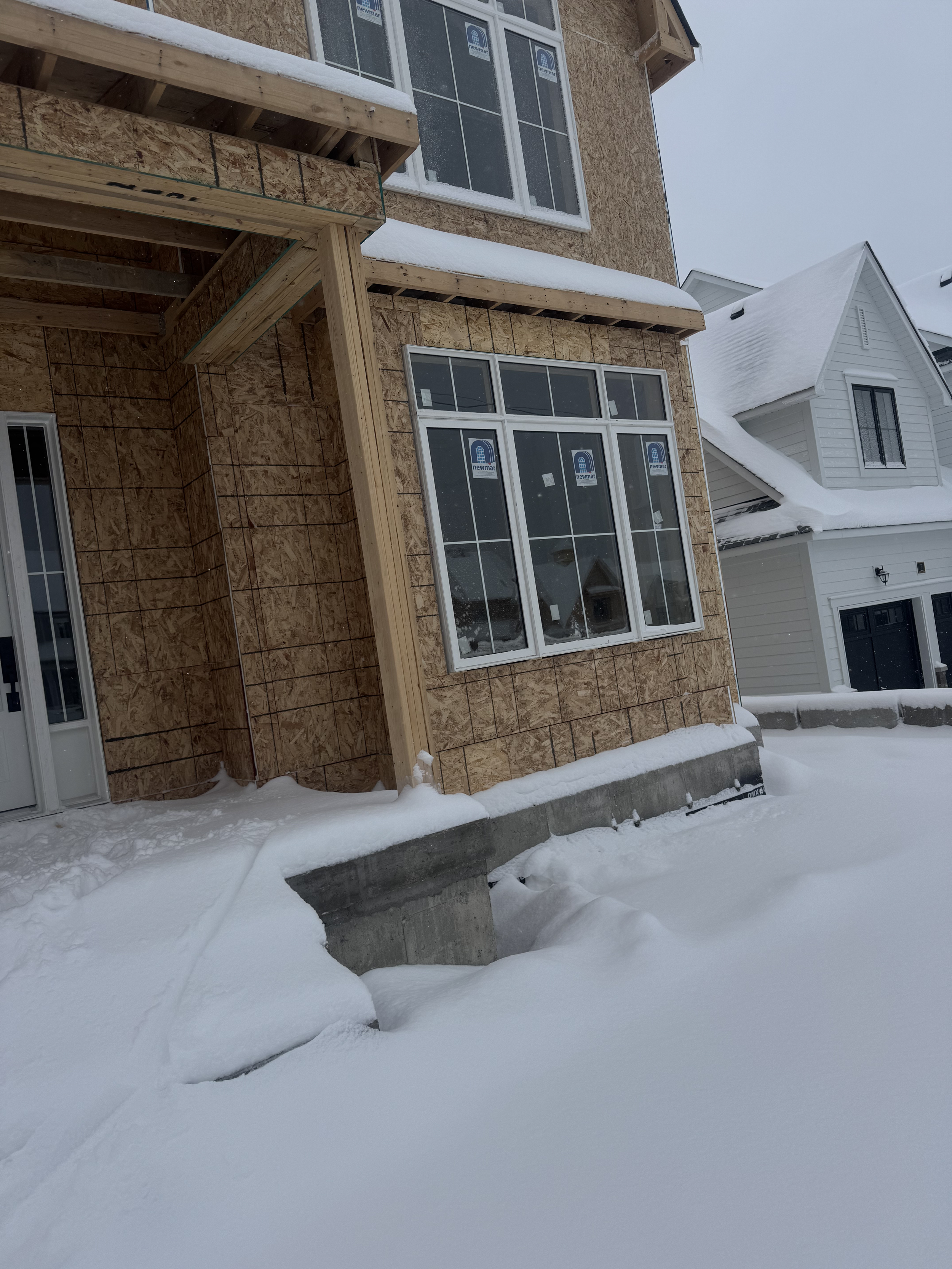 Front view of a house under construction with plywood sheathing, snow on the ground and window frames installed, but no siding yet.