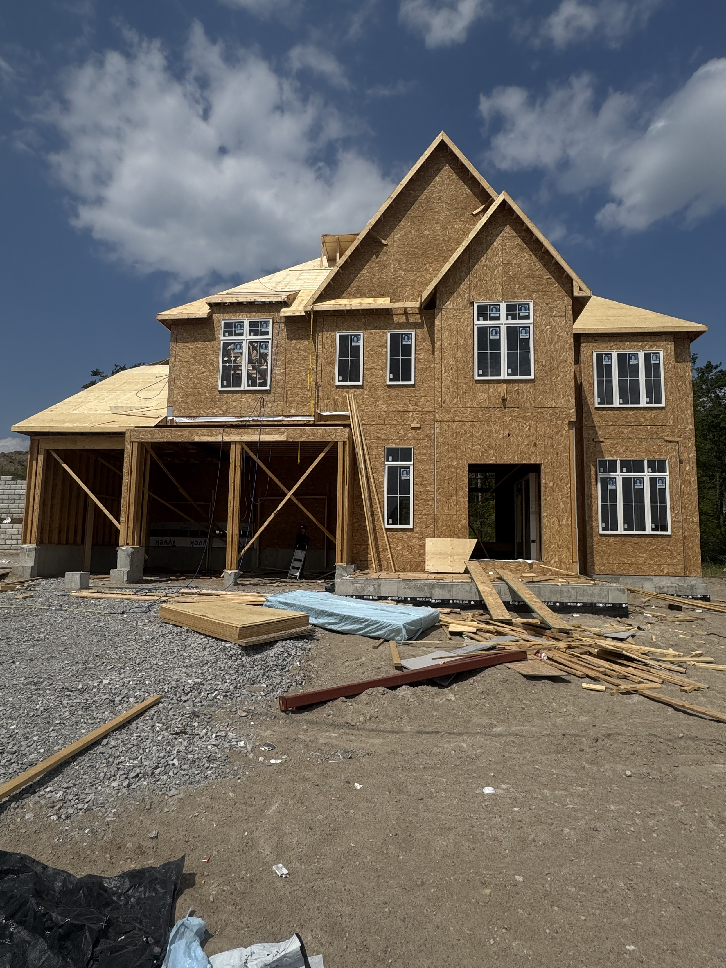 A house under construction with exposed wooden framing and siding, multiple windows installed, and construction materials and debris on the ground.