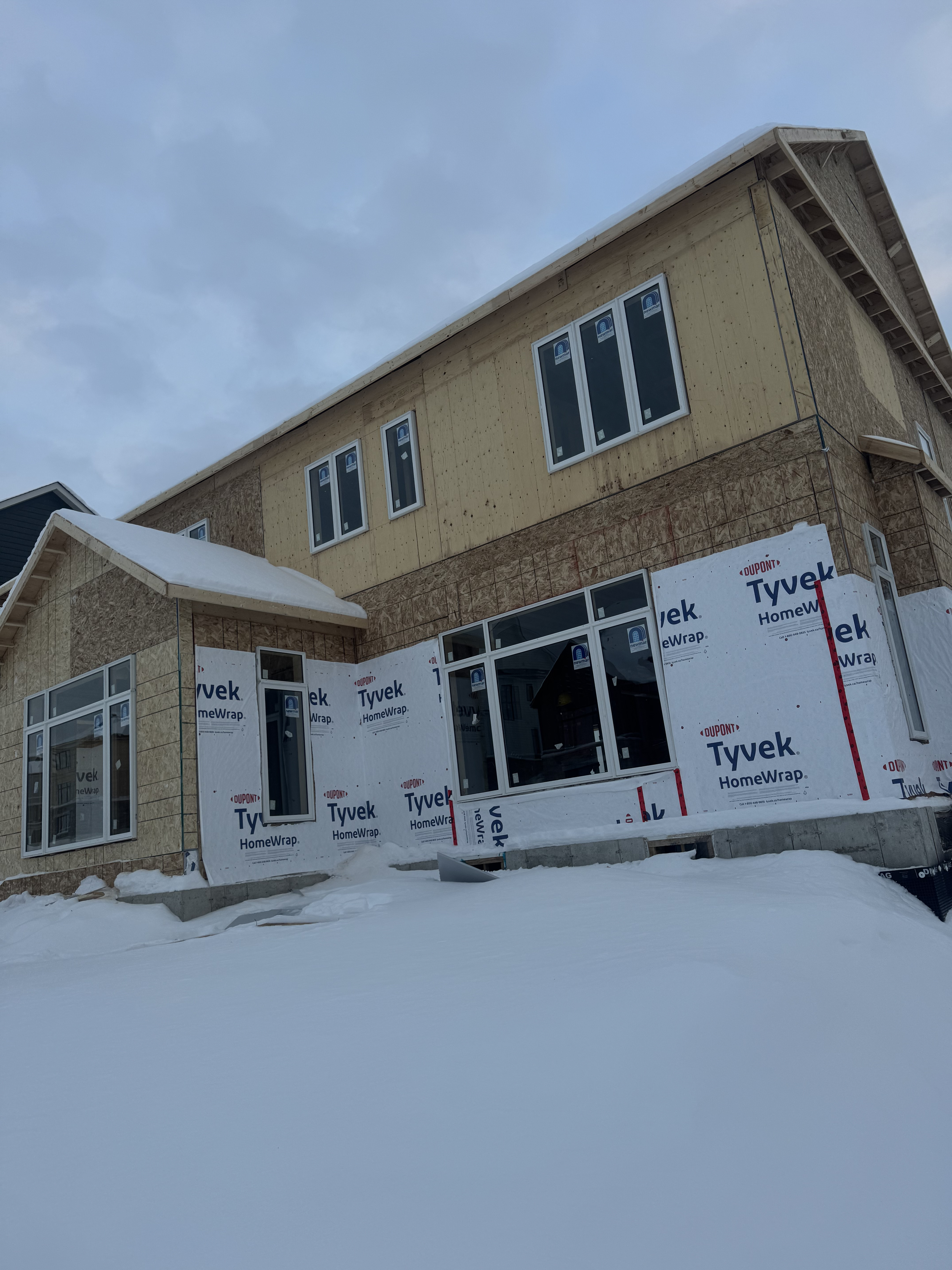 A two-story house under construction with insulation and wrap installed. Snow surrounds the house on the ground.
