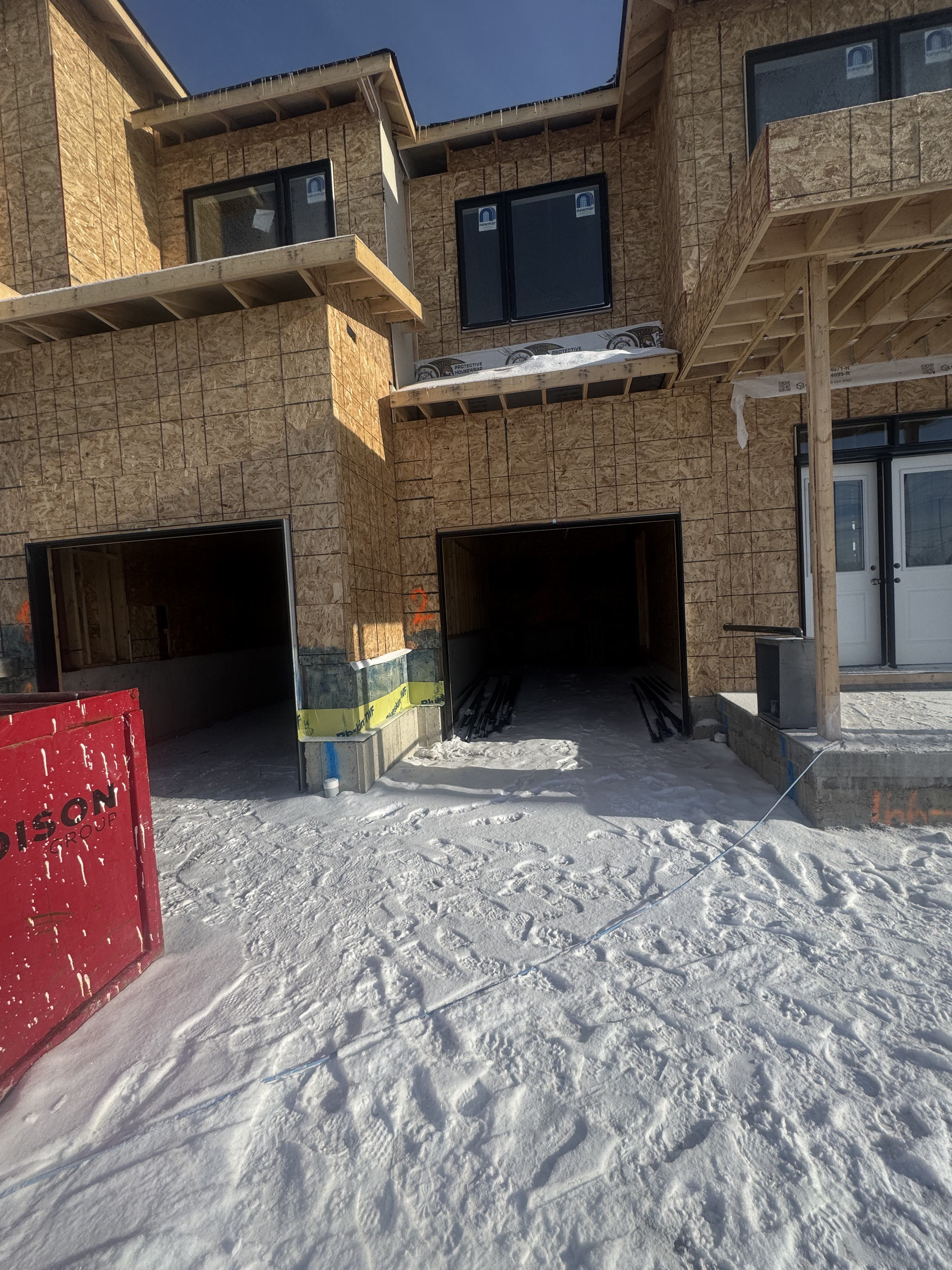 Construction site of a multi-story building with exposed wooden framing and unfinished exterior walls, two open garage spaces, and snow on the ground.