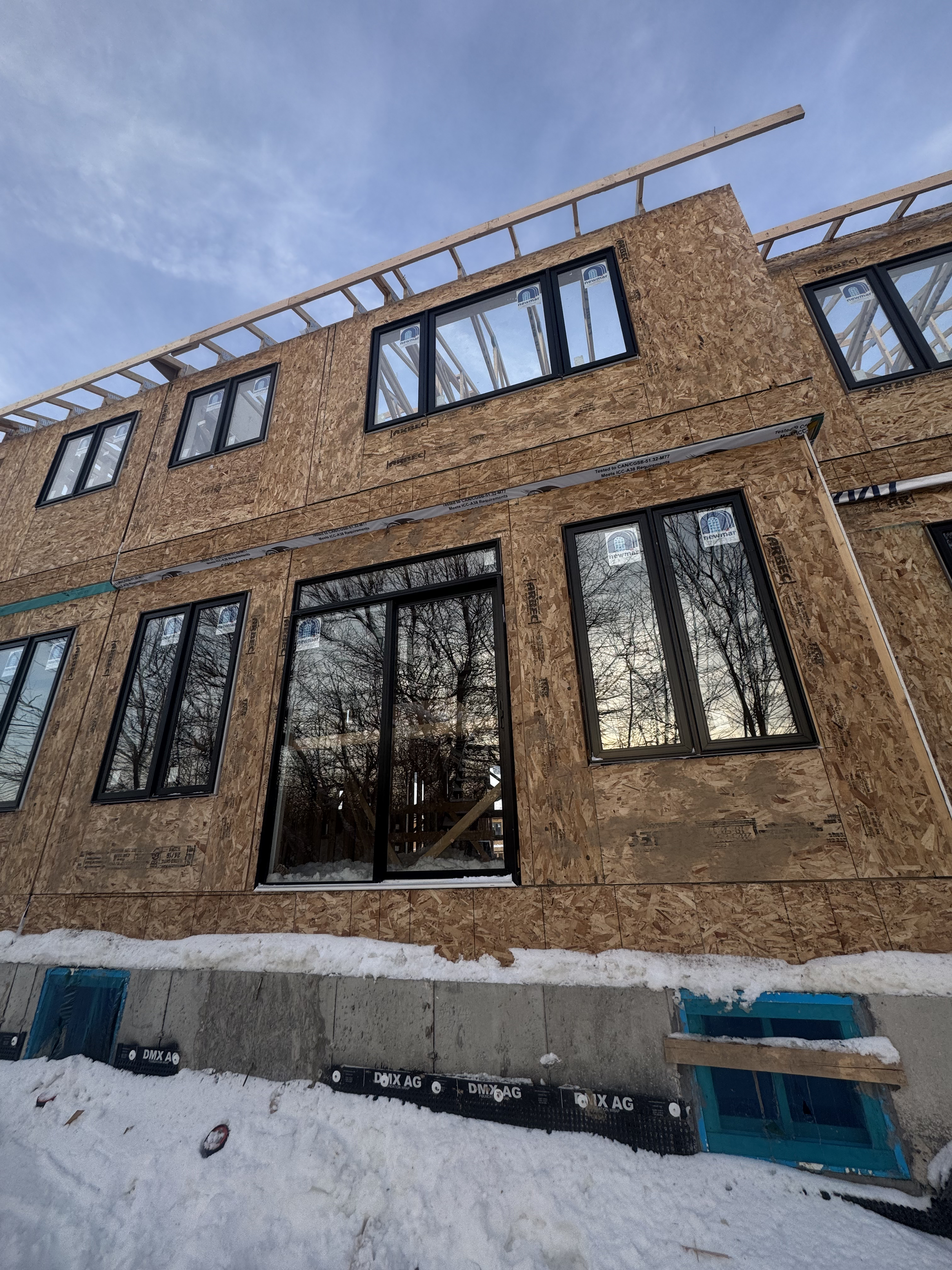 Construction site of a multi-story building with wooden framing and installed windows, surrounded by snow on the ground, under a partly cloudy sky.