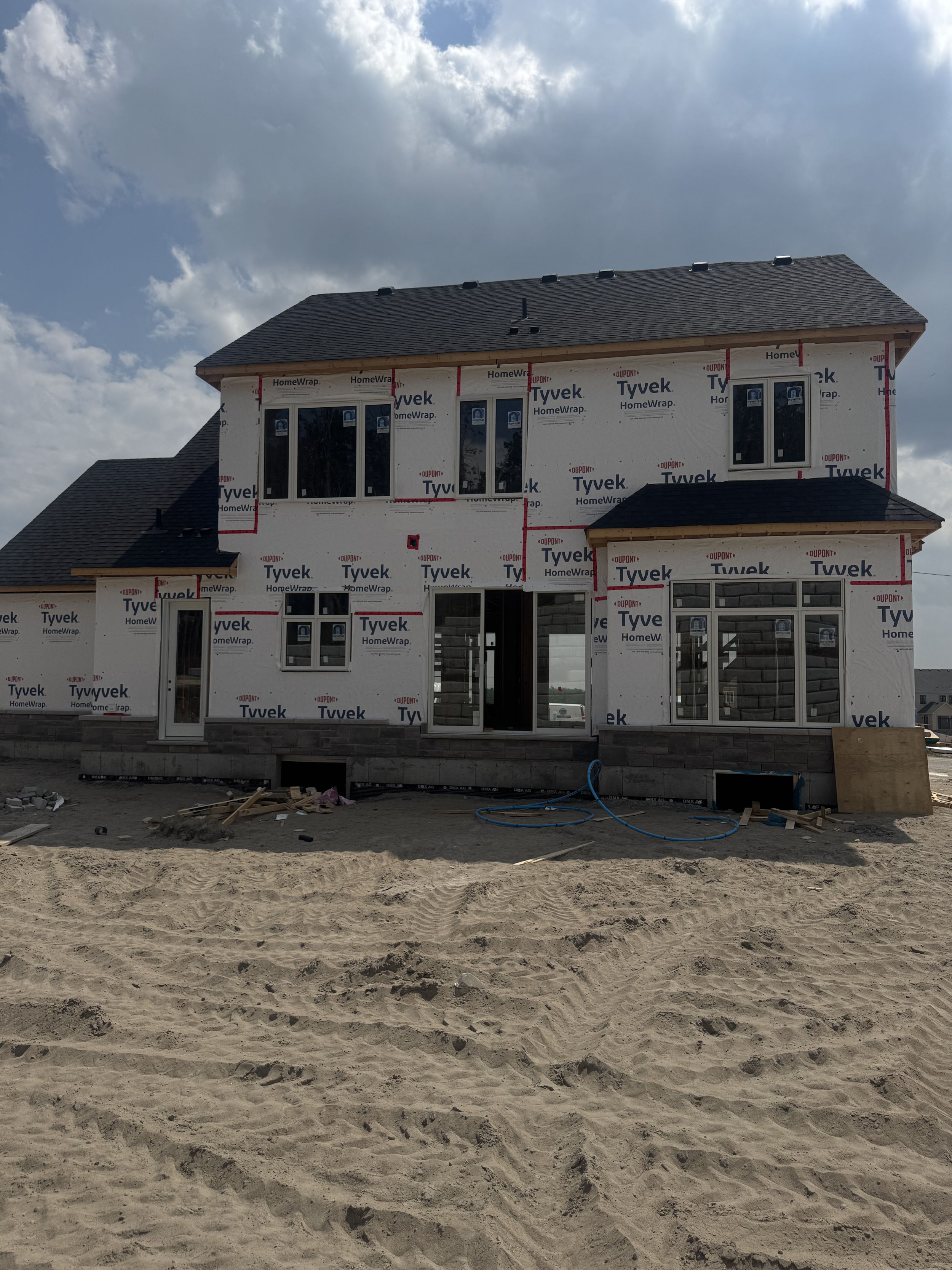 Under construction house with Tyvek HomeWrap siding, multiple windows, and a black shingled roof, situated on sandy ground with construction materials in front.