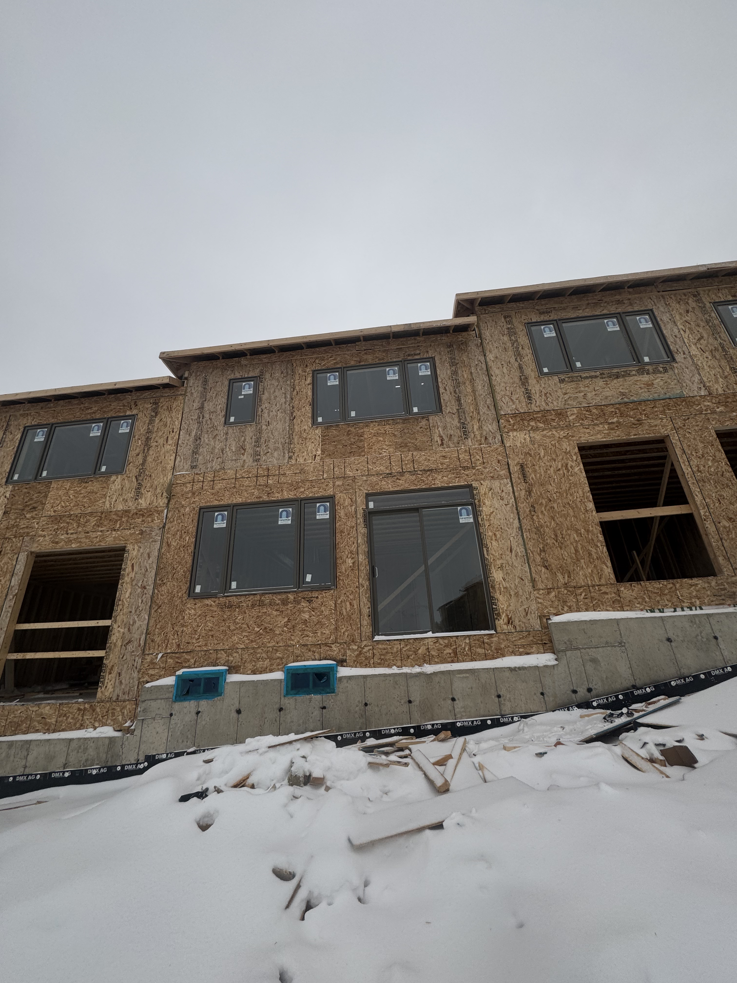 Construction of a multi-story house with unfinished exterior walls and installed windows, snow on the ground, cloudy sky.