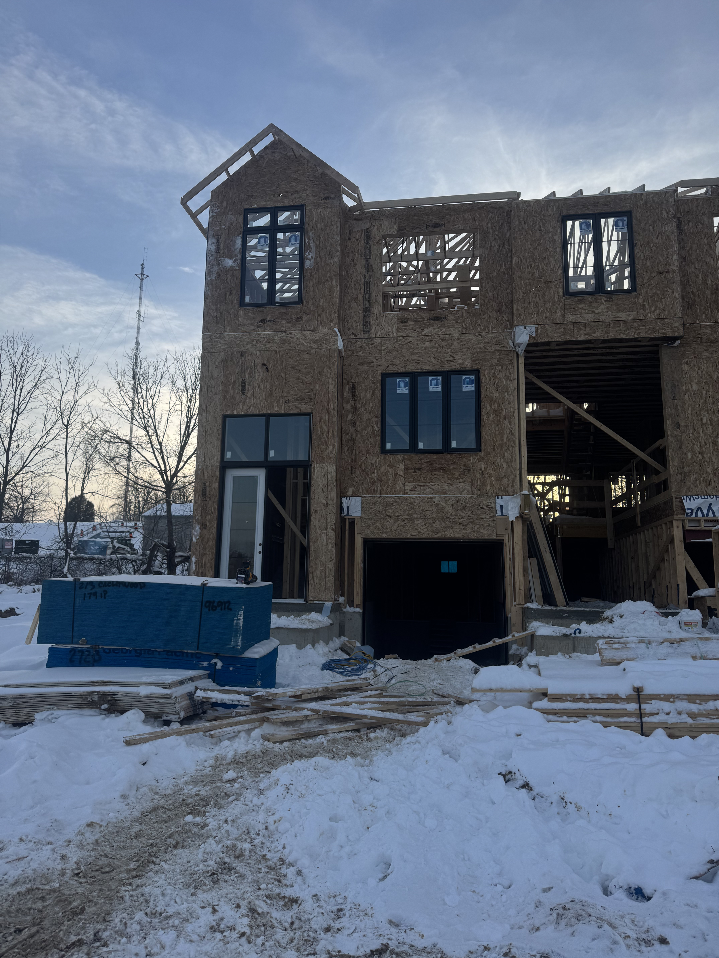 A house under construction with exposed wooden framing, snow on the ground, and a blue sky.