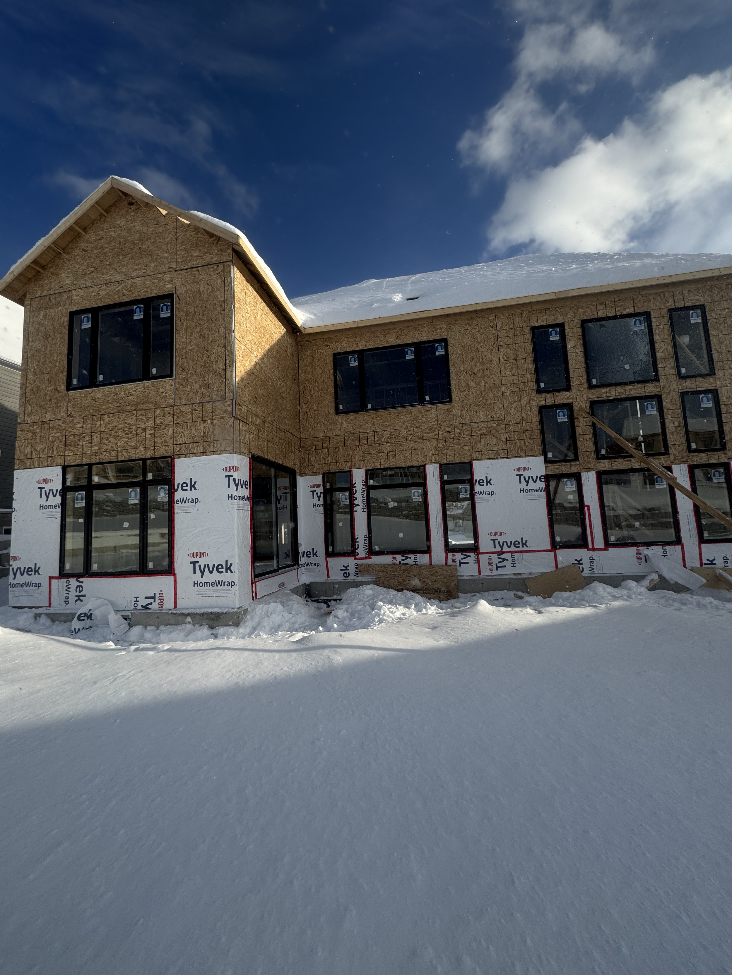 A house under construction with unfinished exterior walls and large window openings, surrounded by snow on a bright winter day.