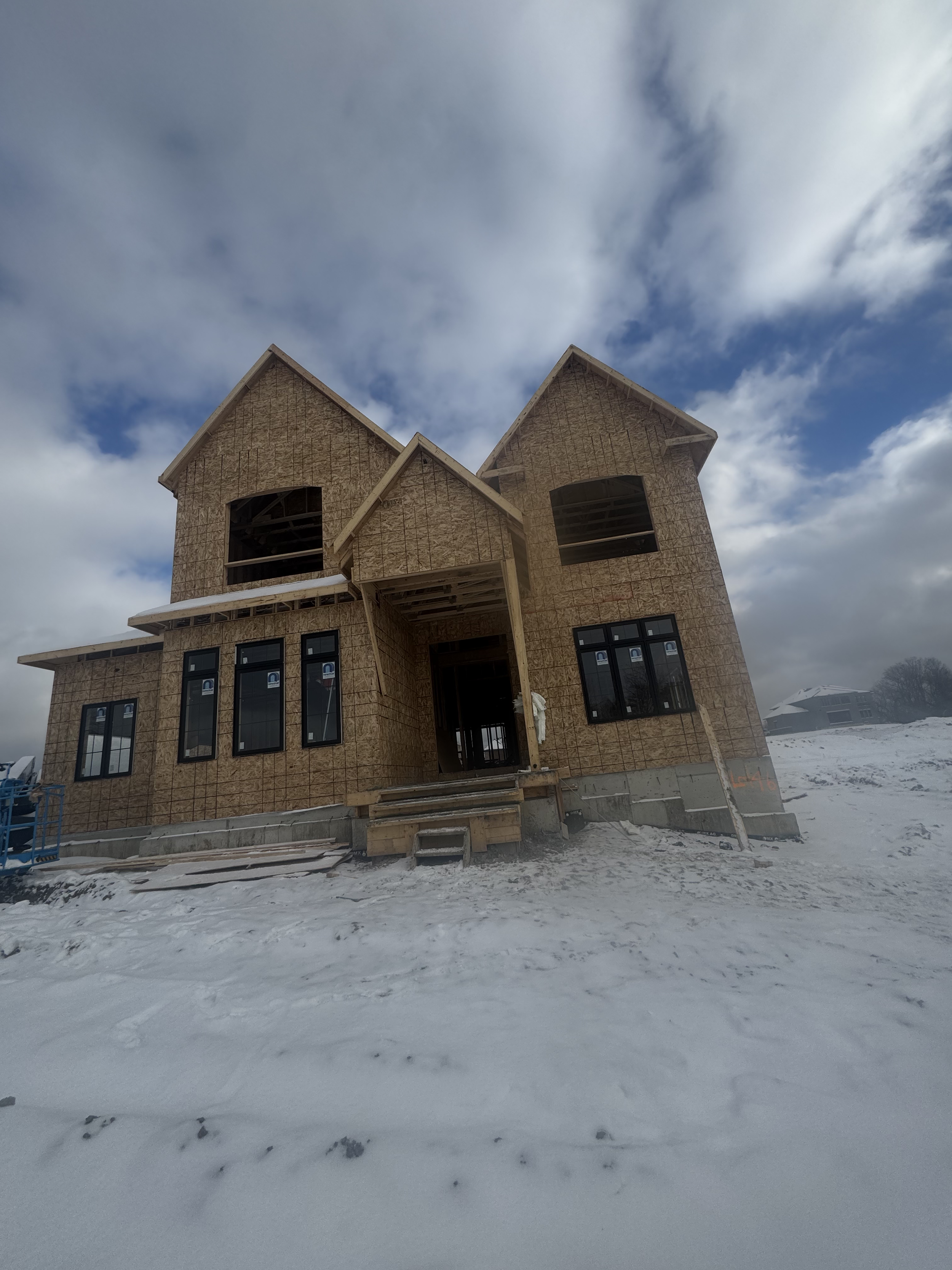 Under construction house with exposed wooden framing, surrounded by snow, against a cloudy sky.