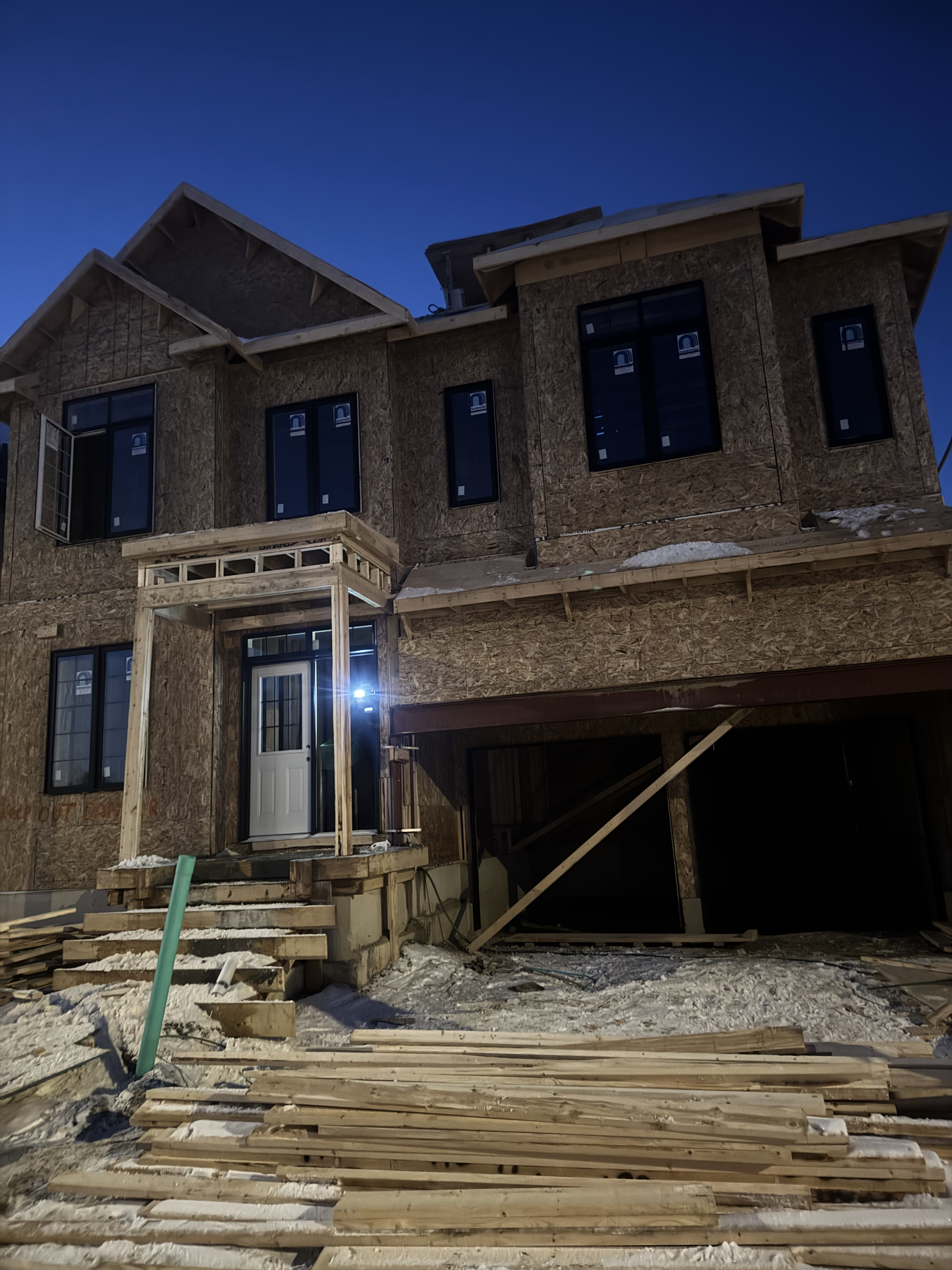 A house under construction with plywood sheathing on the exterior walls, a front door with a porch, and several window openings. Construction materials and debris are on the ground, and the scene is illuminated by an artificial light during evening o