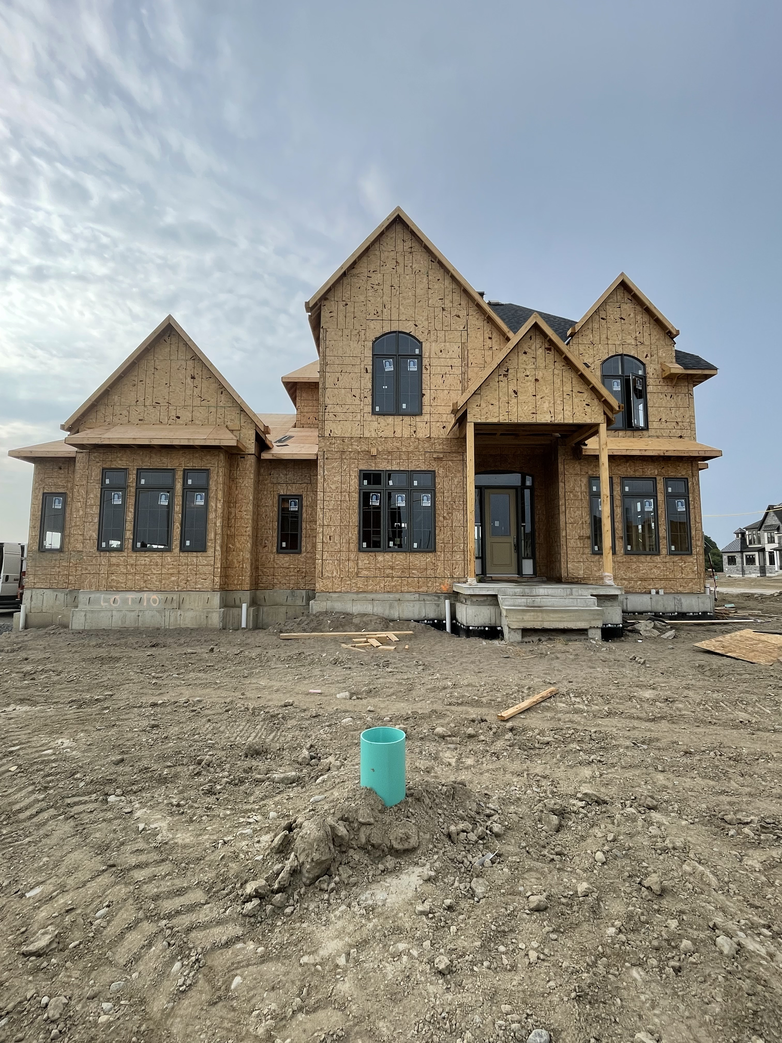 A two-story house under construction with unfinished exterior walls and many windows, situated on a dirt lot with construction materials around.