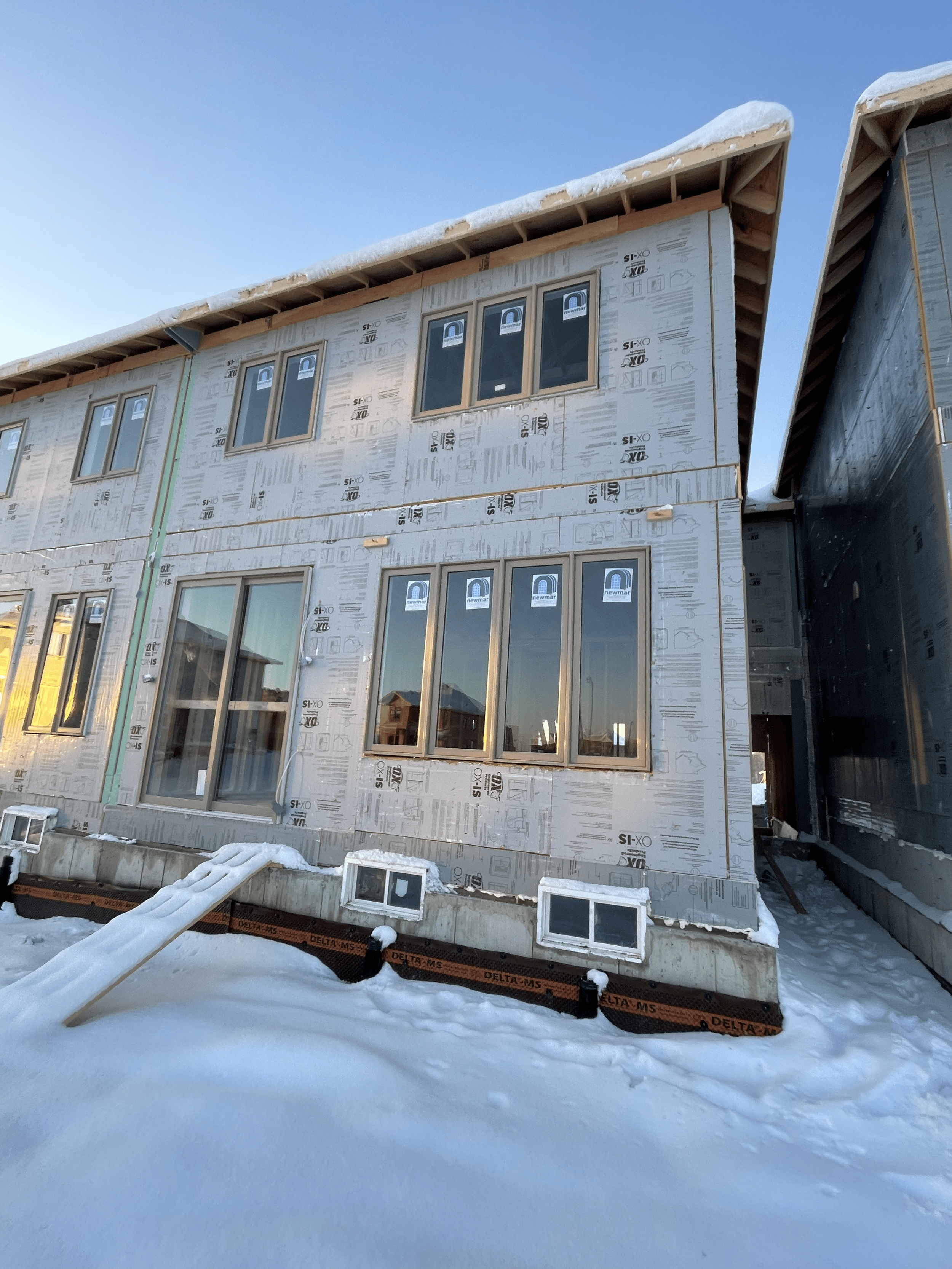 Newly constructed multi-story house under construction with snow on the ground, several windows installed, and a blue sky.