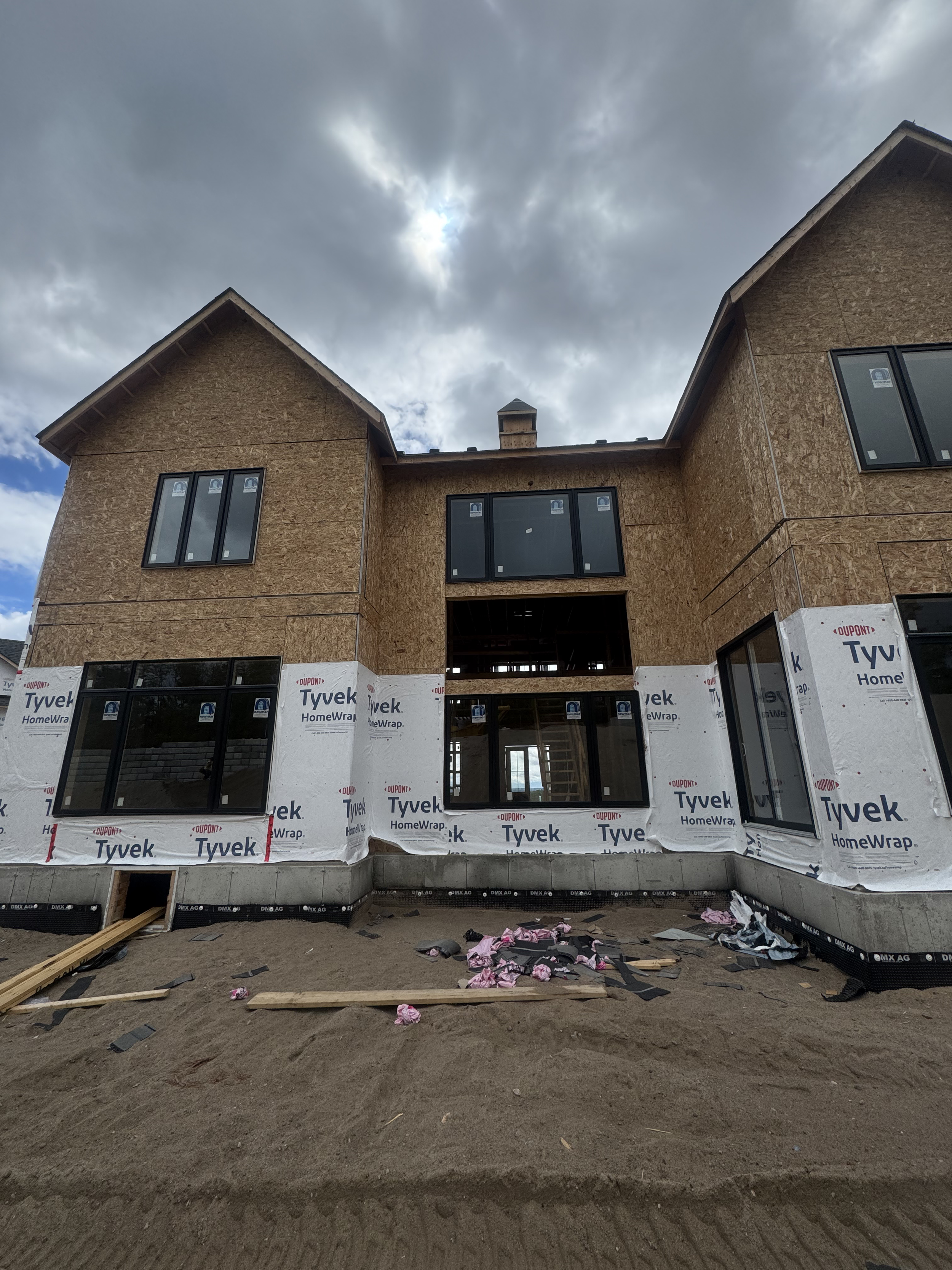 A house under construction with unfinished exterior walls, wrapped in Tyvek HomeWrap, and large window openings without glass installed, surrounded by construction debris and dirt on a cloudy day.