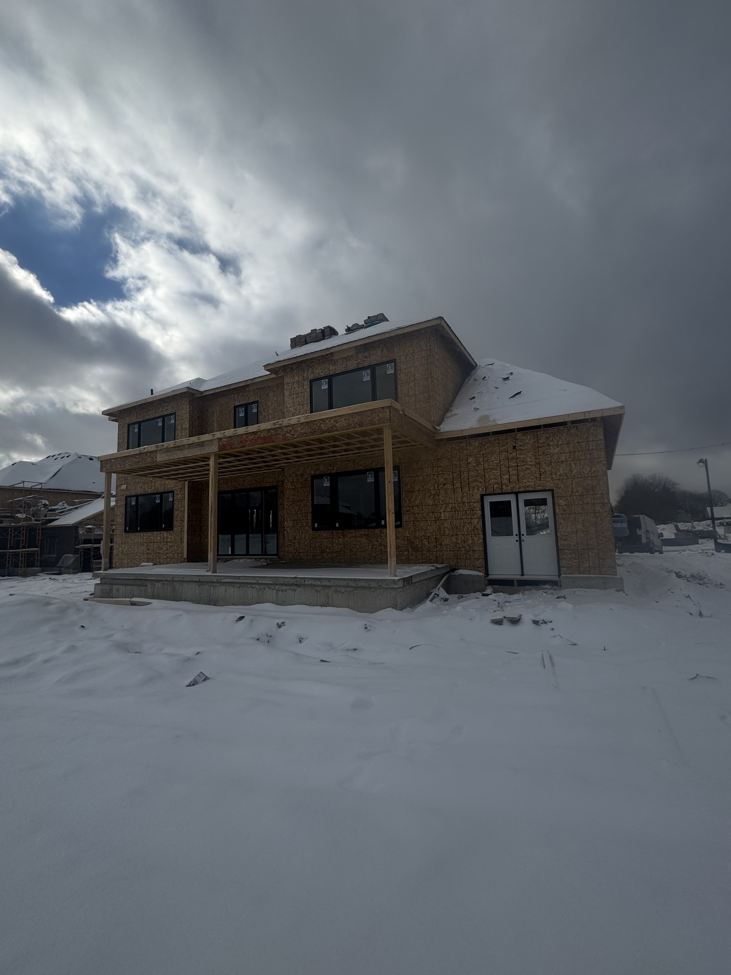 A house under construction with wooden framing, large windows, and a snow-covered roof, in a snowy landscape.