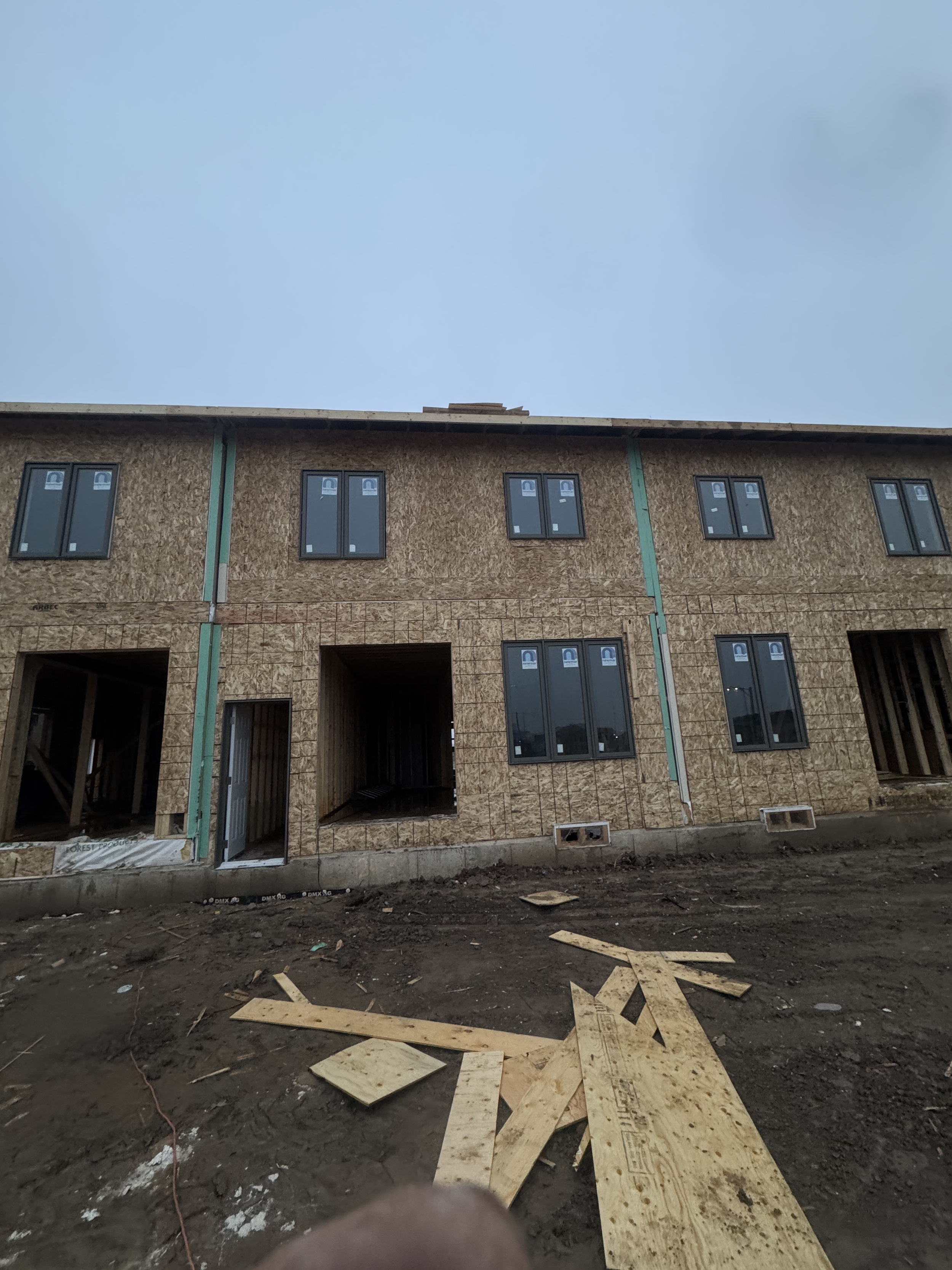 Under-construction multi-story building with unfinished wood framing and black-framed windows, construction debris, and dirt ground in the foreground.