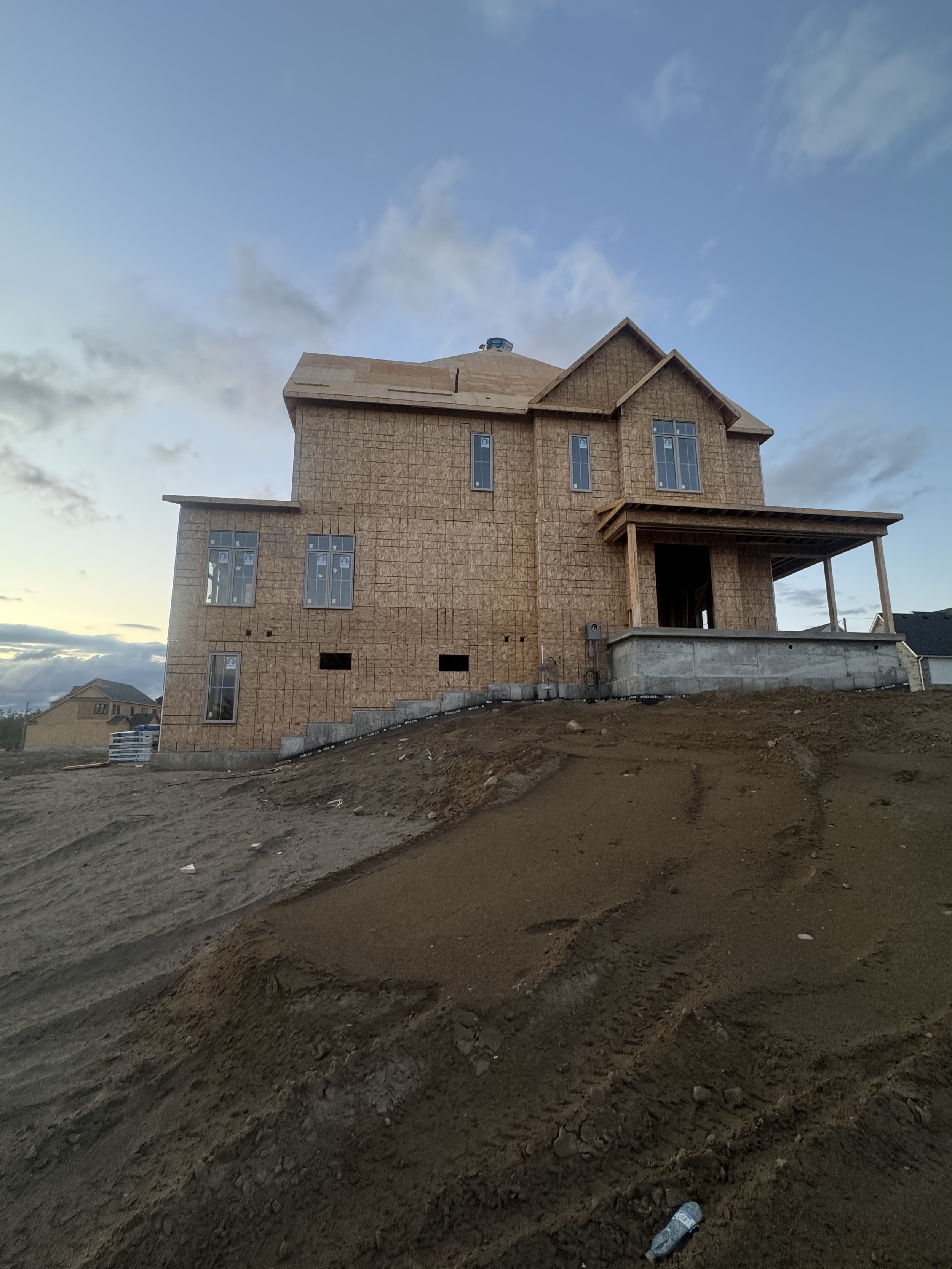 A house under construction on a dirt hill, with exposed wooden framing and windows, set against a sky with scattered clouds.