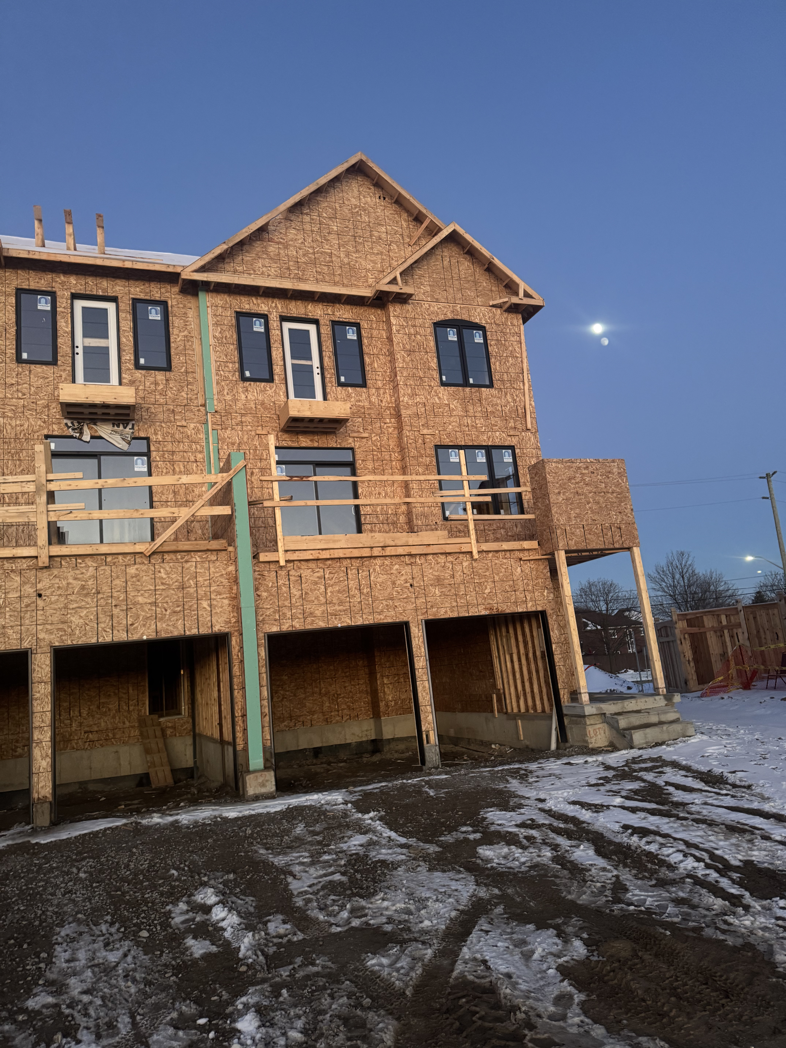 A multi-story house under construction with wooden framing and black window frames, set against a clear sky with visible moon, and snow on the ground.