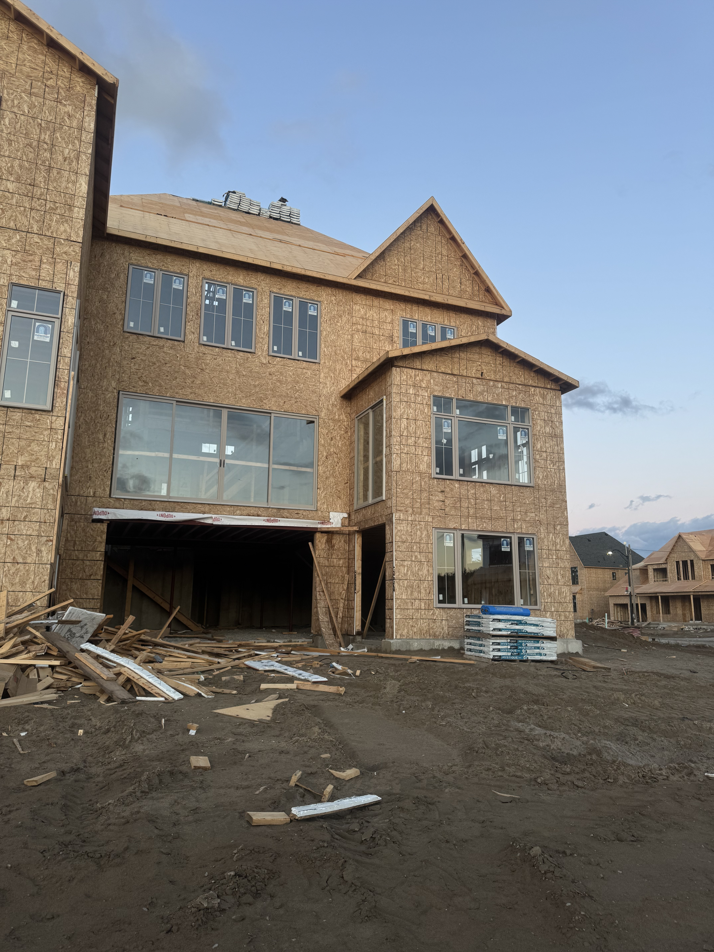 Under-construction multi-story house with exposed wooden framing, large windows, and unfinished exterior, surrounded by dirt and construction materials.