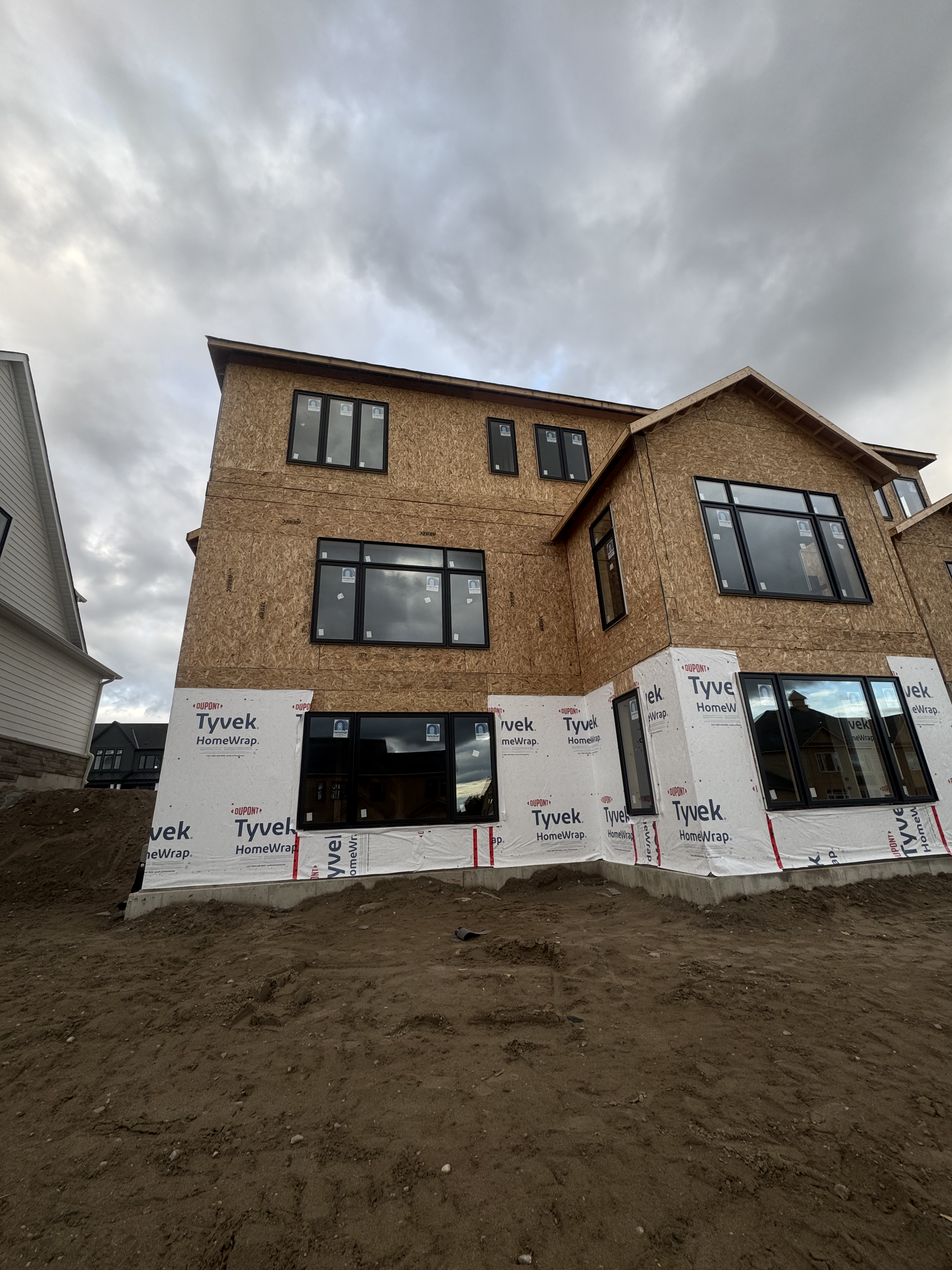 A multi-story house under construction with exposed sheathing and new windows installed, on a dirt lot with cloudy sky above.