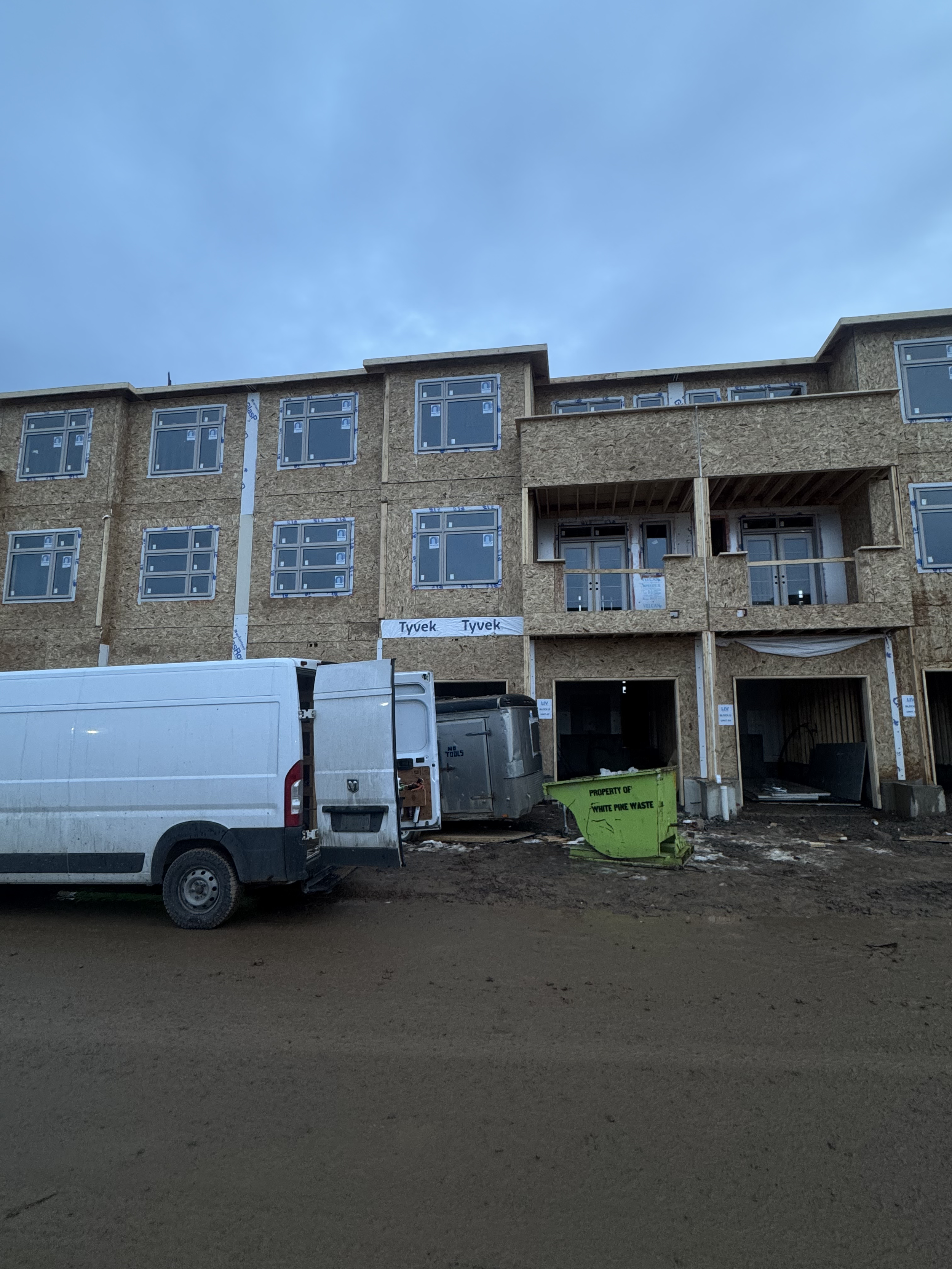 Under-construction apartment building with wooden framing, windows installed, construction vehicles, and a green waste dumpster in front