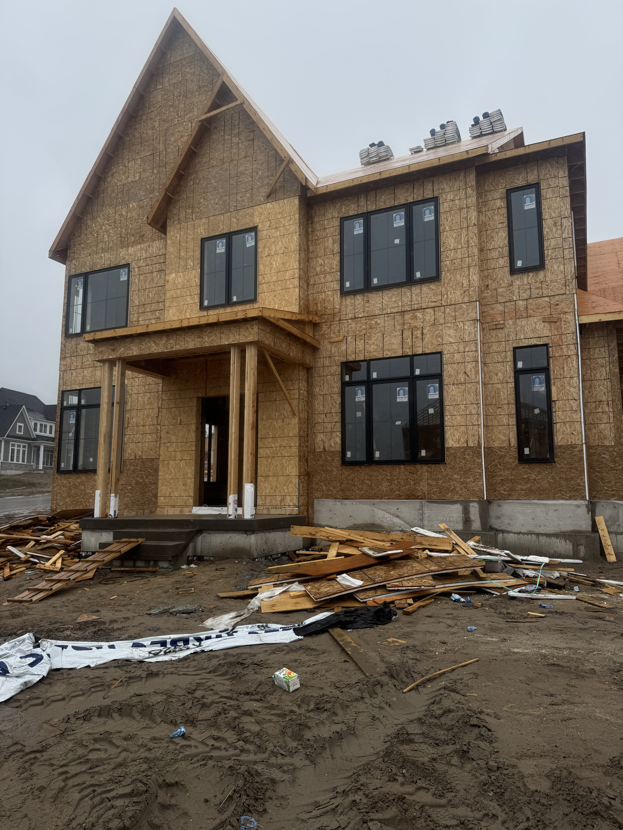 Under construction two-story house with exposed plywood exterior and black window frames, surrounded by dirt and construction debris.