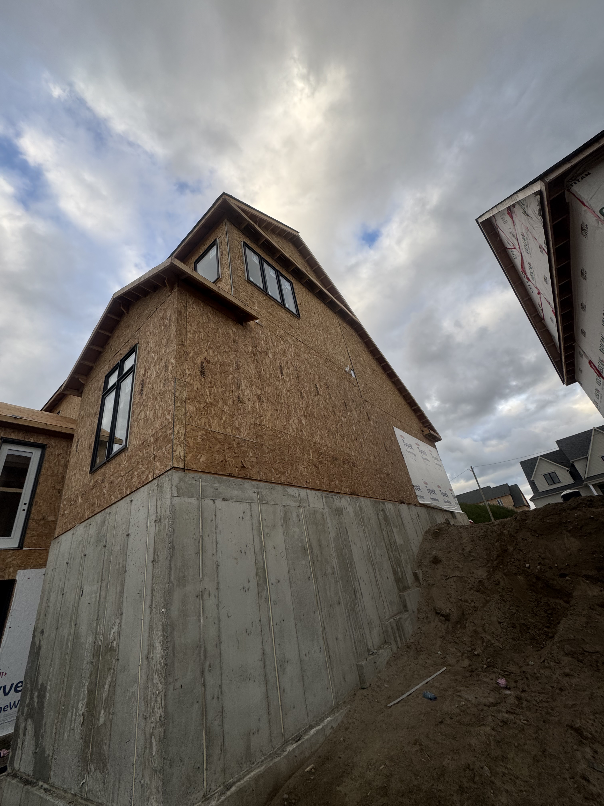 A house under construction with wooden sheathing on its exterior walls, a concrete foundation, and a partially visible neighboring house. The sky is cloudy with patches of blue.