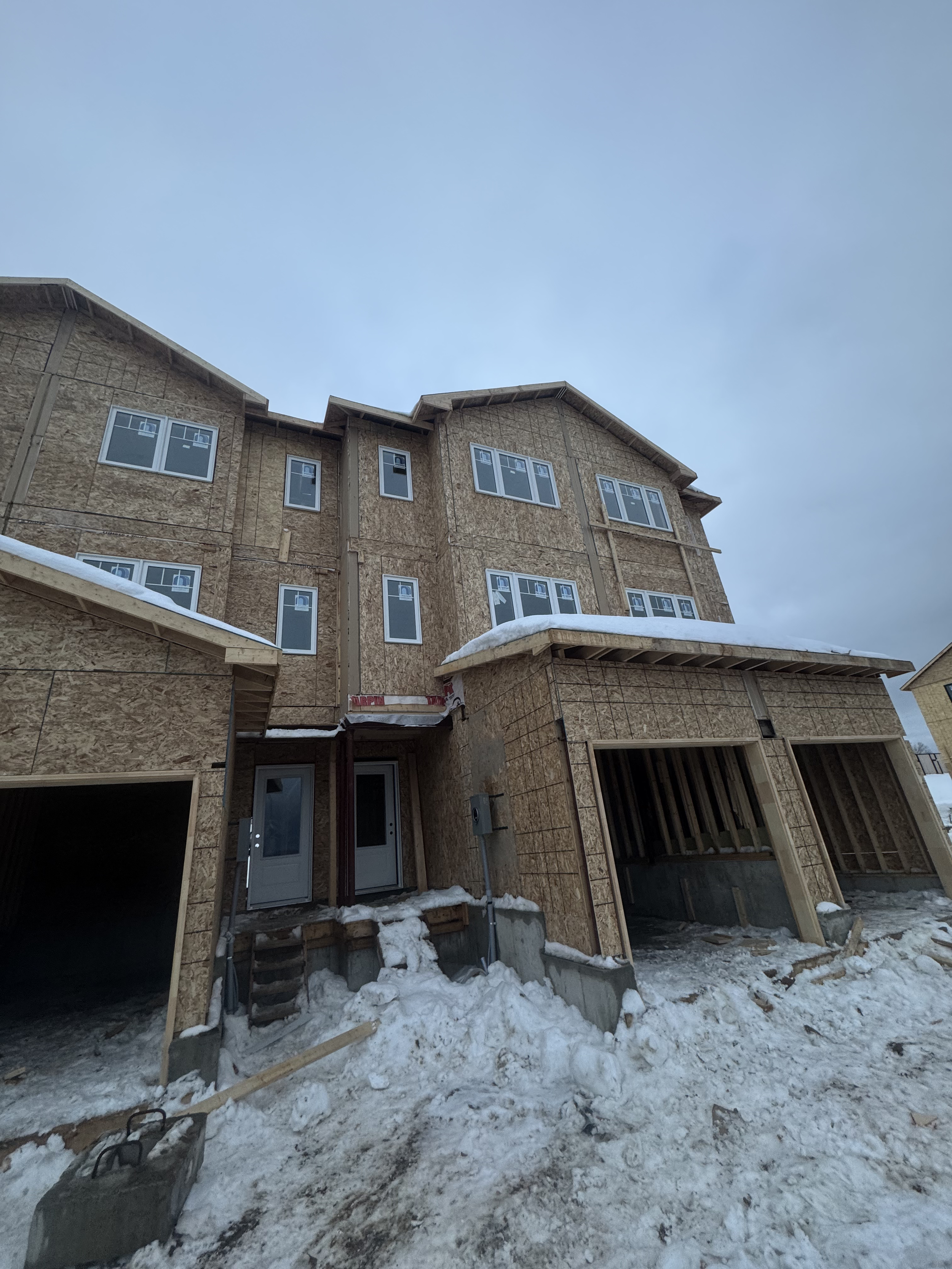 Under-construction multi-story house with exposed wooden framing, snow on the ground, and cloudy sky.