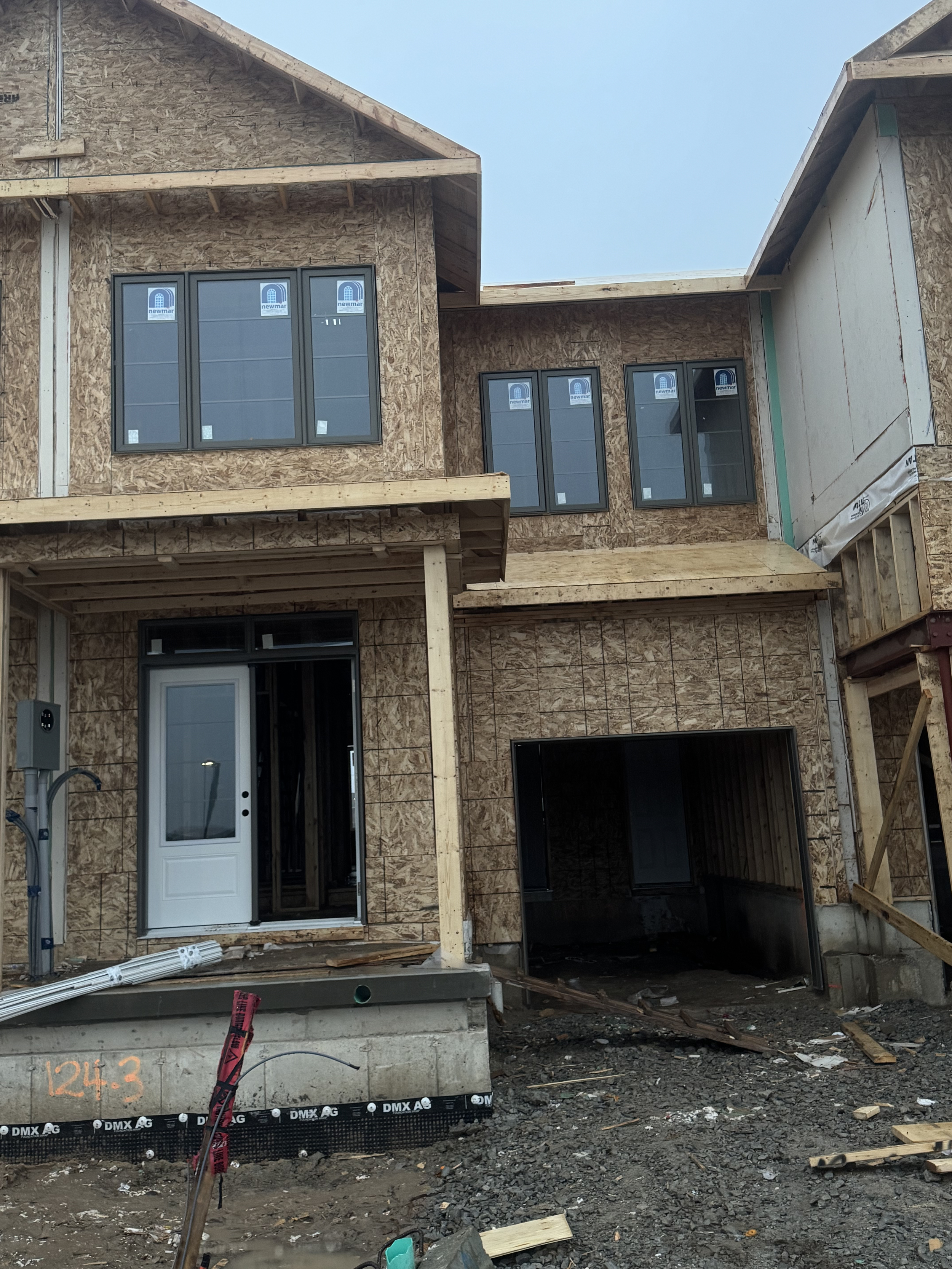 A multi-story house under construction with OSB sheathing and new windows installed, featuring an open garage and front porch area.
