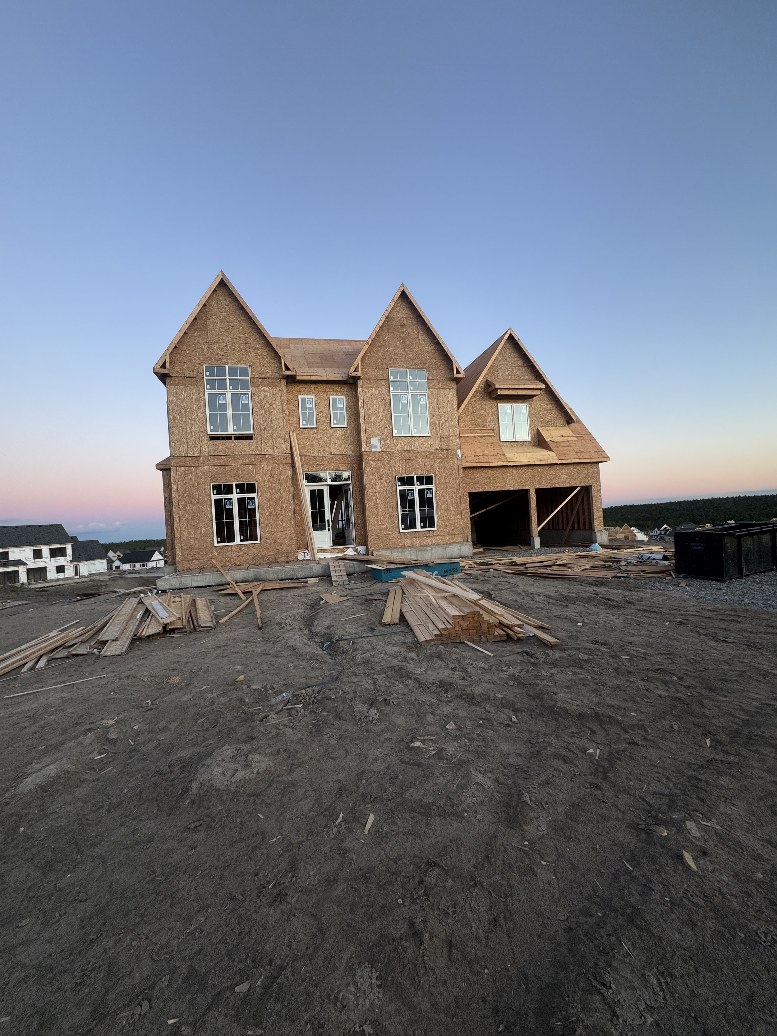 A house under construction with exposed wooden framing, showing multiple windows, set against a sunset sky, with construction materials on the ground.