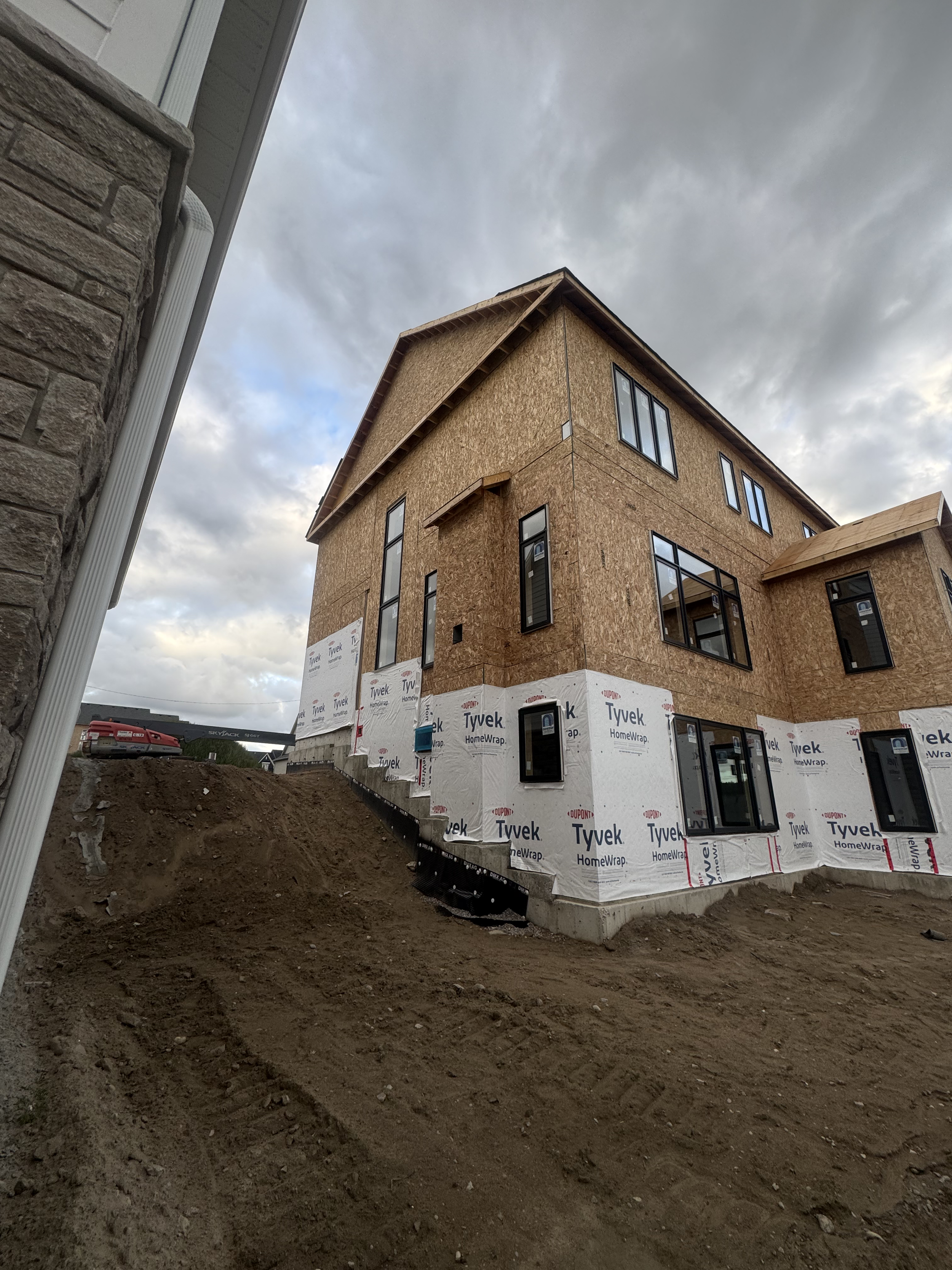 A multi-story house under construction with a wood frame exterior, several windows, and a concrete foundation. The house is surrounded by dirt.
