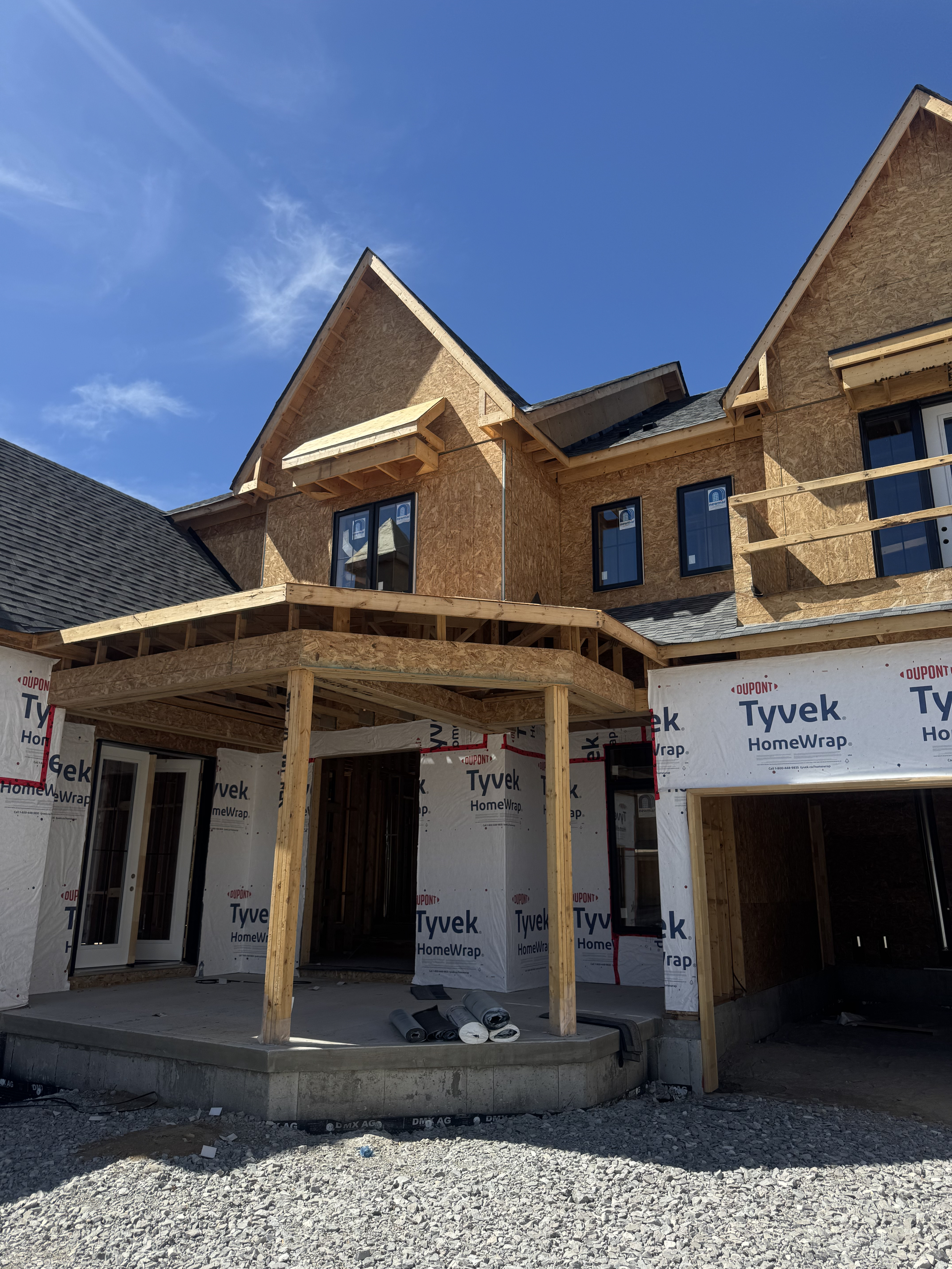 A multi-story house under construction with exposed wood framing, black framed windows, and partial siding covered in Tyvek HomeWrap. The sky is clear and blue.