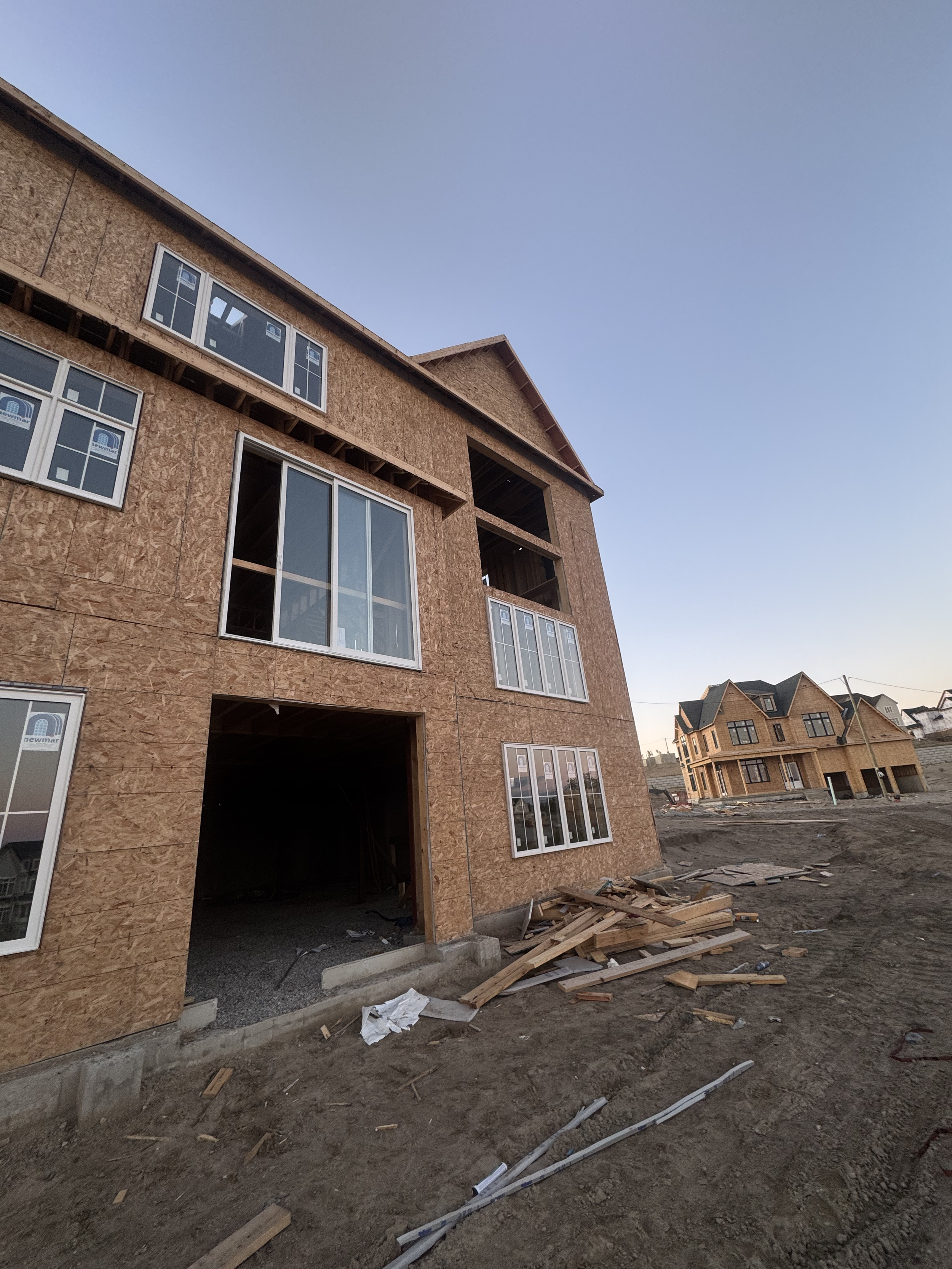 Under construction residential building with plywood exterior, multiple large windows, and an open garage entryway. Construction materials and debris on the ground in front of the building.