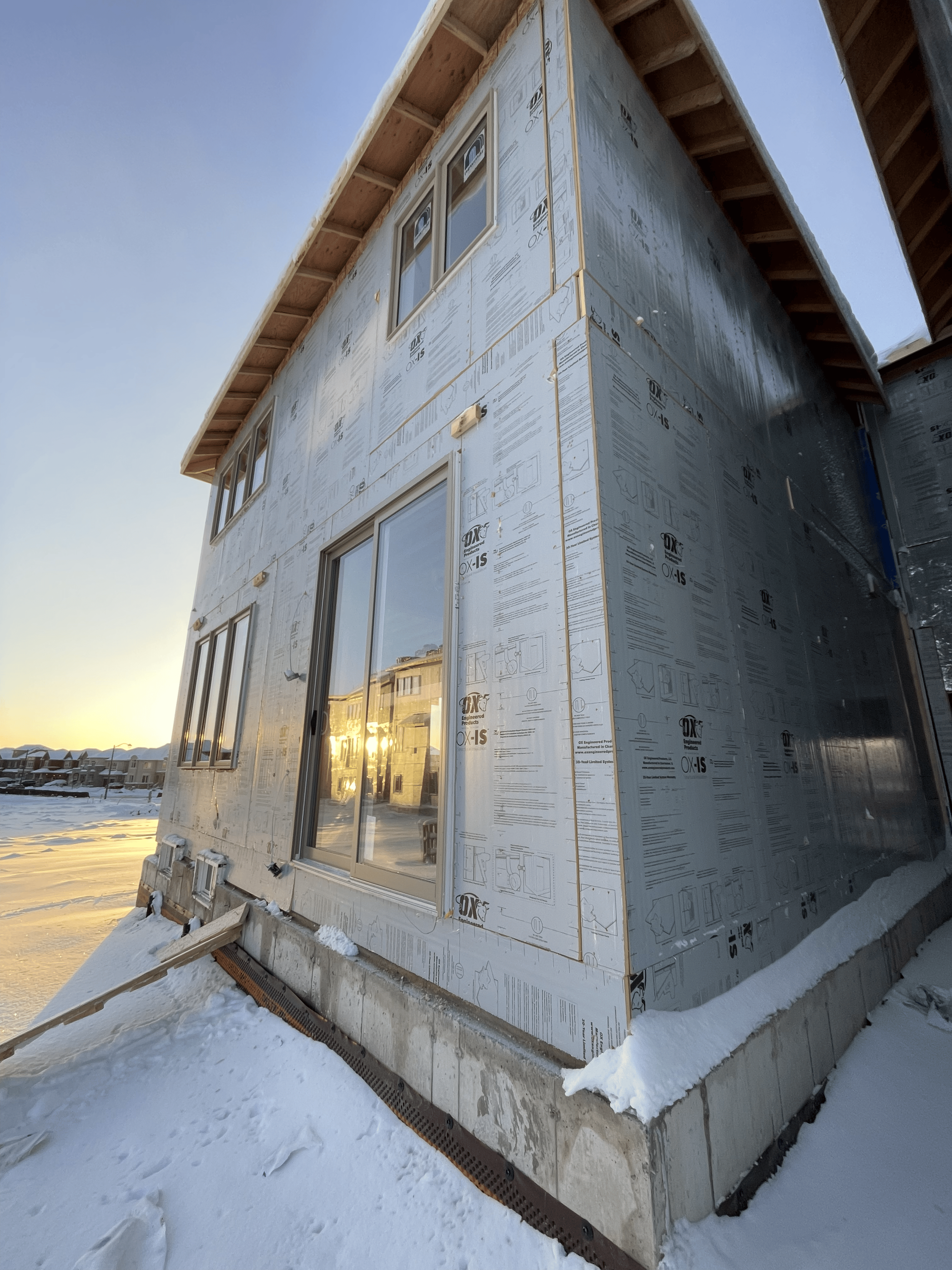Side view of a house under construction with exterior sheathing, windows, and snow on the ground at sunset.