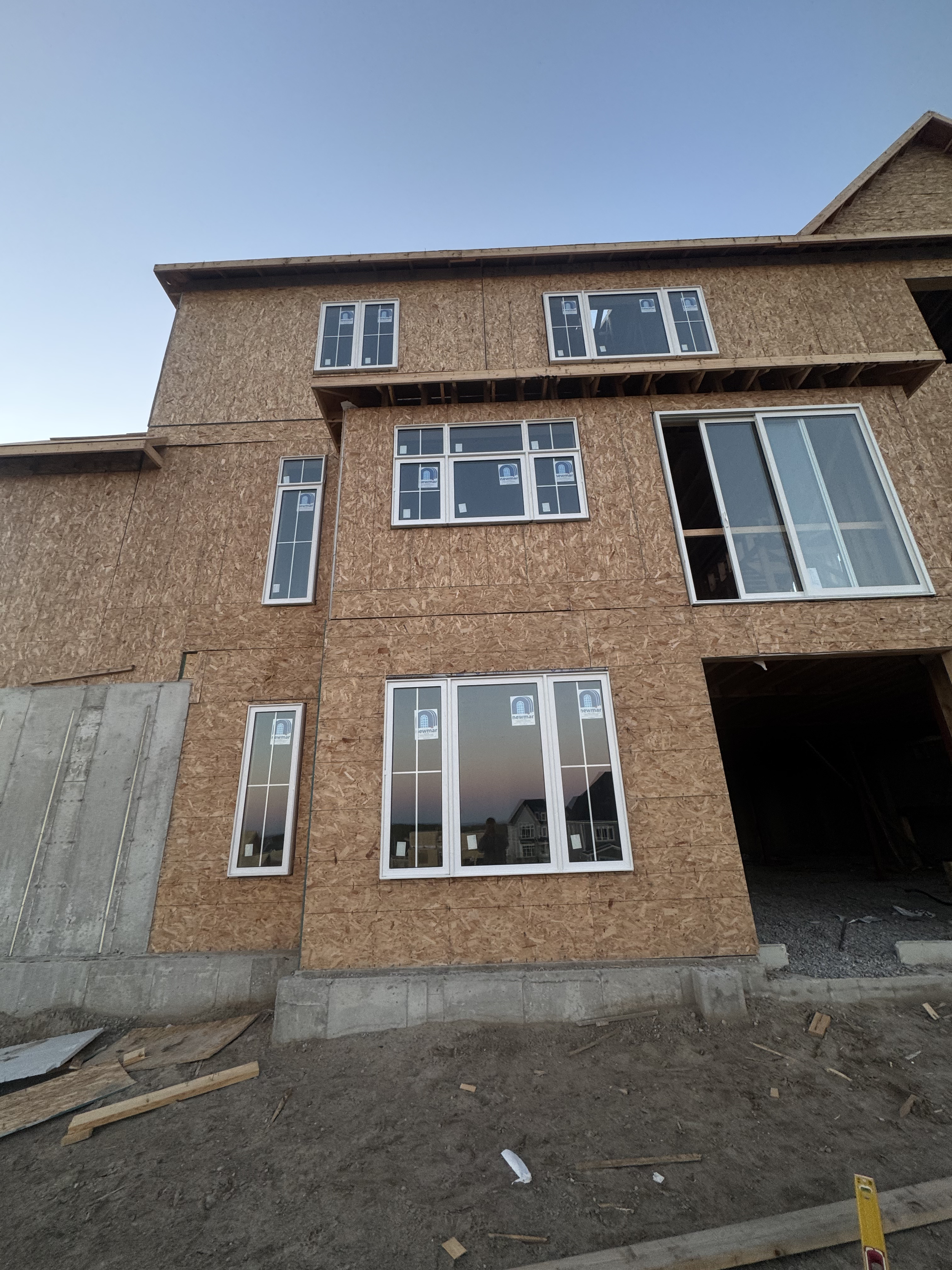 A multi-story house under construction with plywood walls and installed windows, with some exterior groundwork in the foreground.