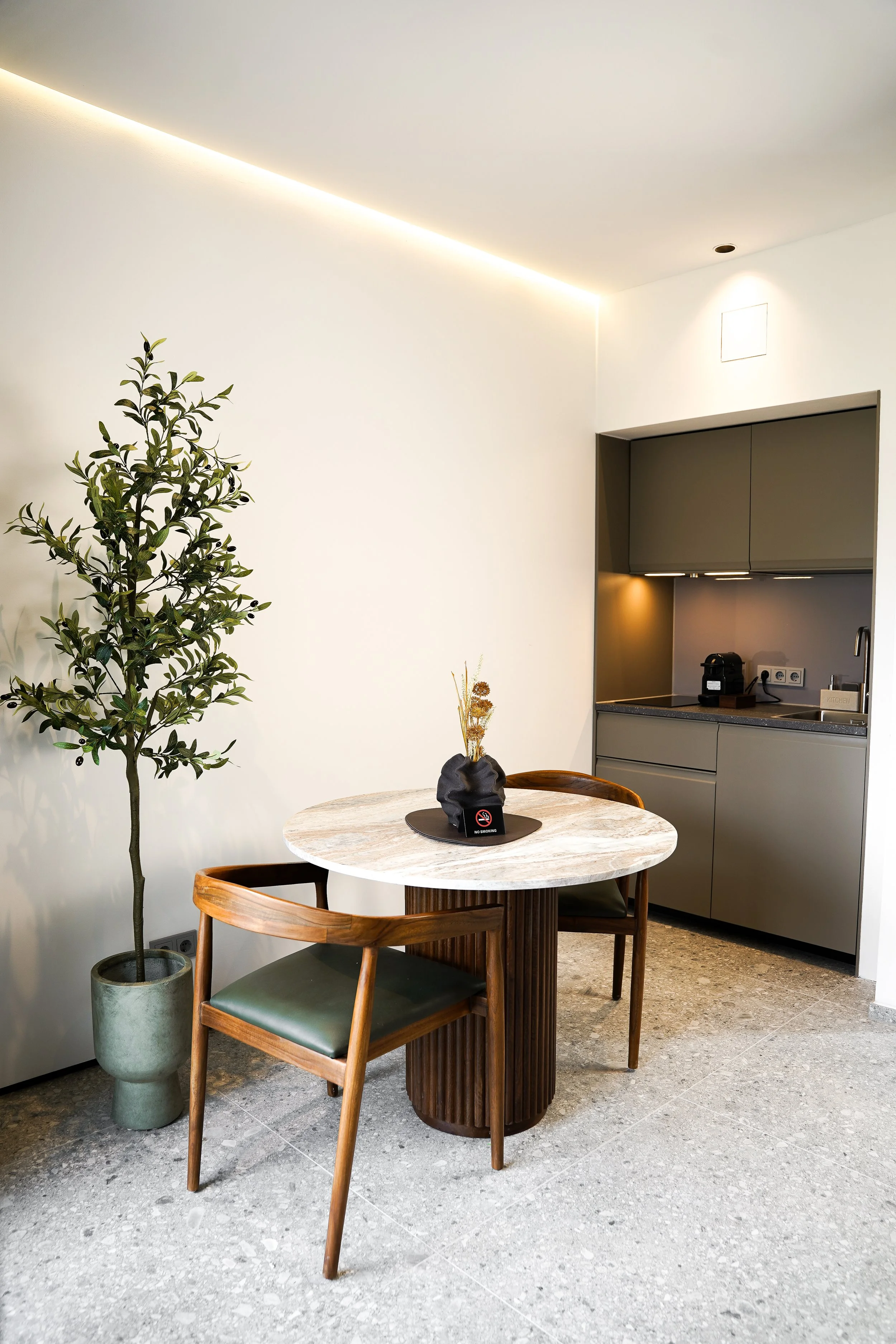 Kleine Küchenecke mit grauem Schrank, runder Marmortisch, zwei Holzstühlen mit grüner Sitzfläche, Topfpflanze mit großen Blättern, und dekorativer Vase mit Trockenblumen auf dem Tisch.