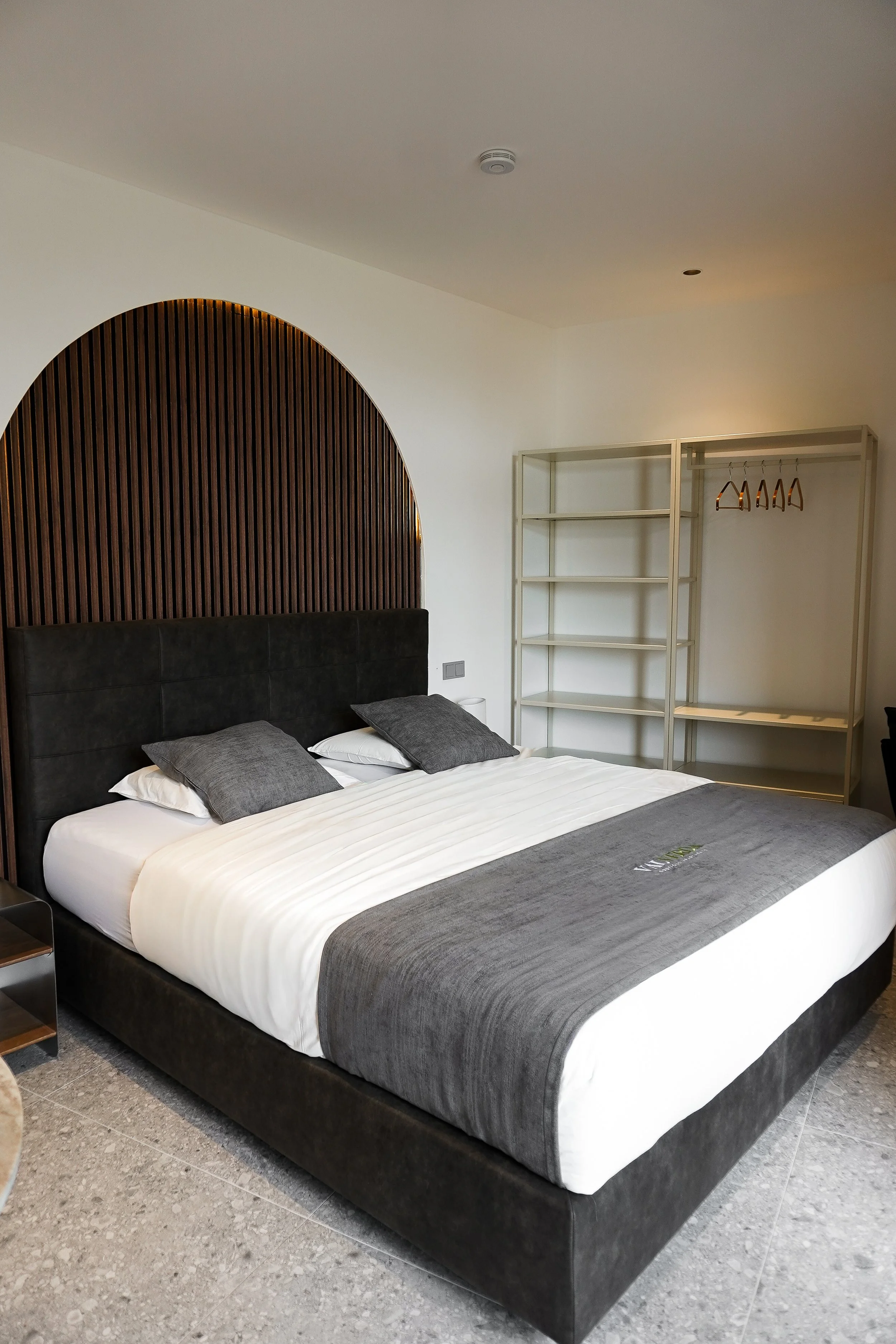 Modernes Schlafzimmer mit schwarzem Bett, weißen und grauen Bettwaren, offener Kleiderschrank und hellen Wänden.