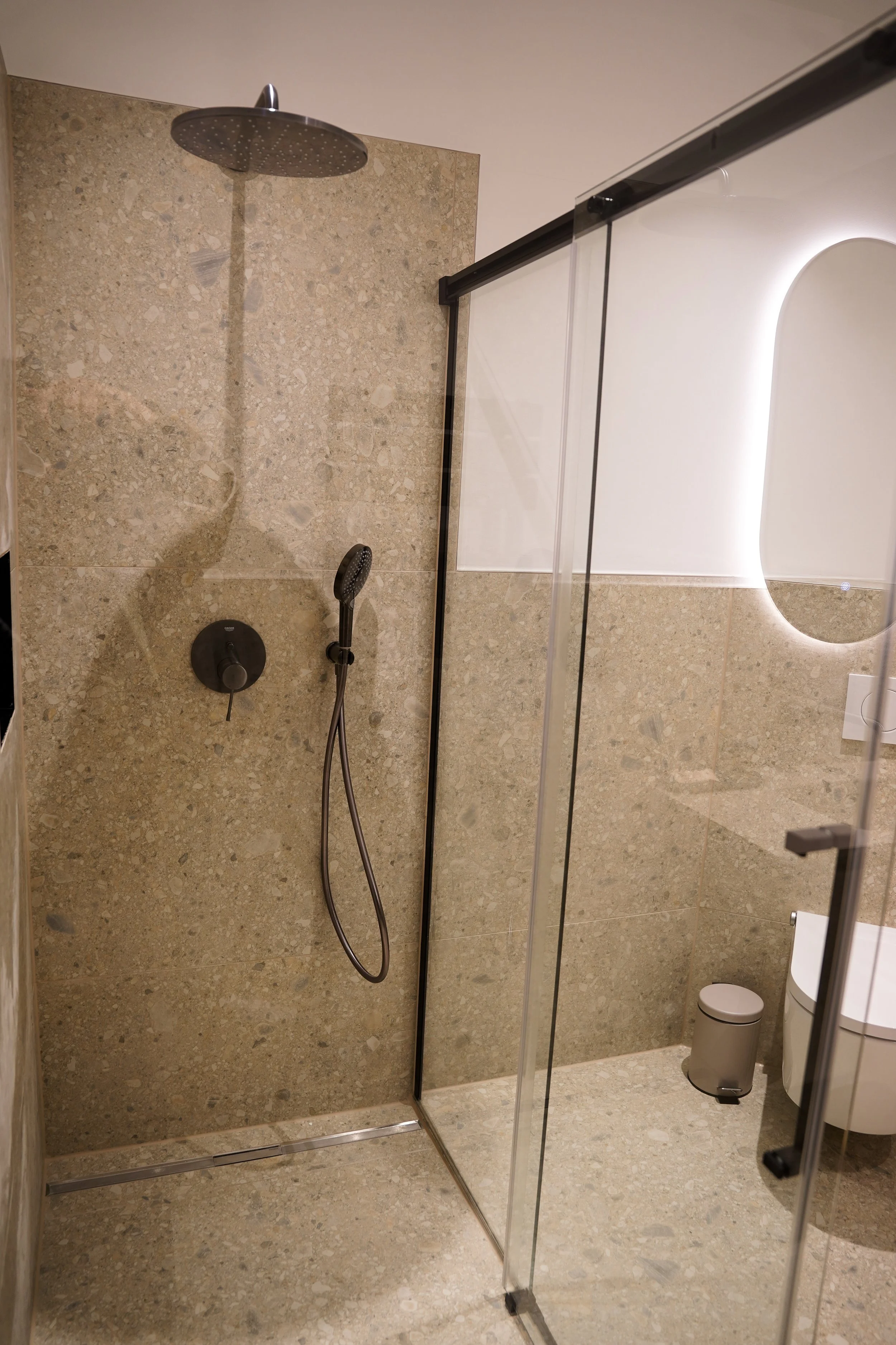 Moderner Badezimmerbereich mit begehbarer Dusche, grauer Steinwand, runden Handbrause und einem kleinen Mülleimer neben einem weißen WC. Spiegel mit Beleuchtung im Hintergrund.