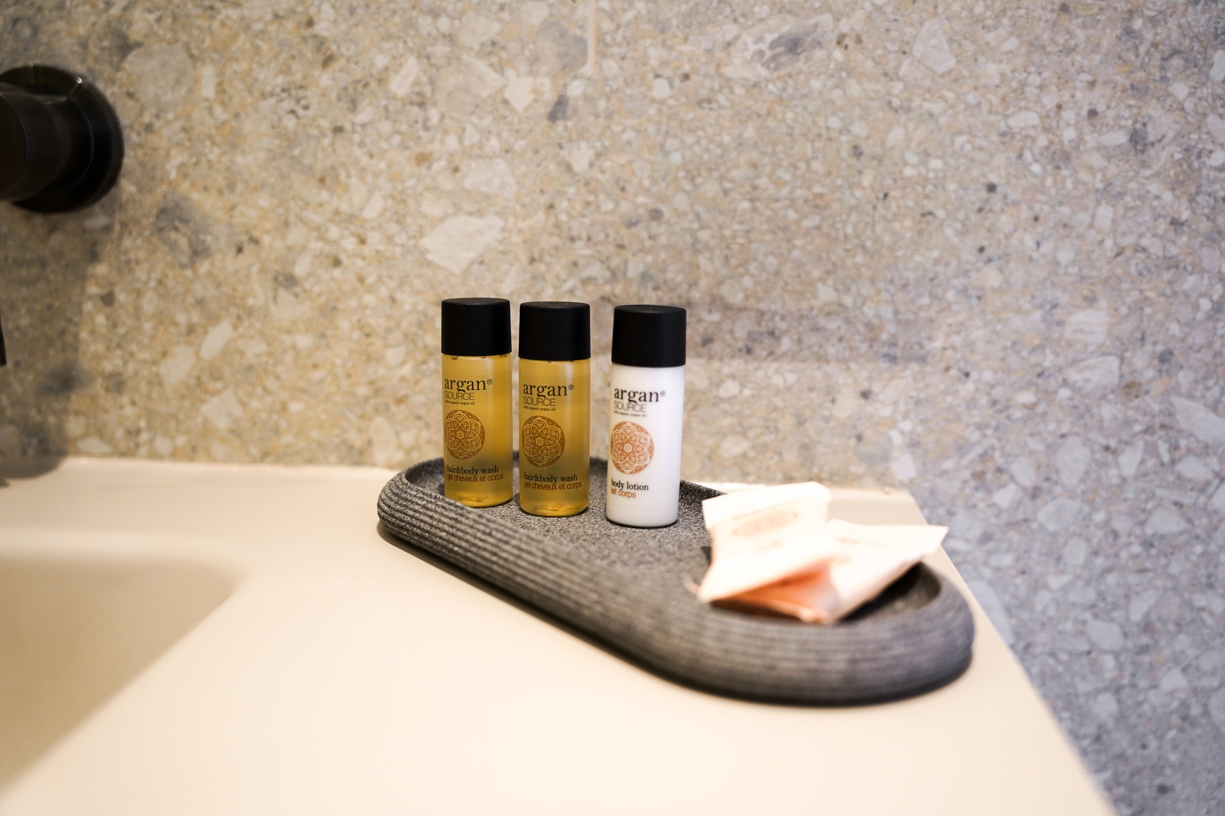 Drei kleine Flaschen mit Kosmetikprodukten, zwei mit gelbem Inhalt und eins mit weißem, auf einer dunklen Ablage in einem Badezimmer, neben einem Waschlappen.