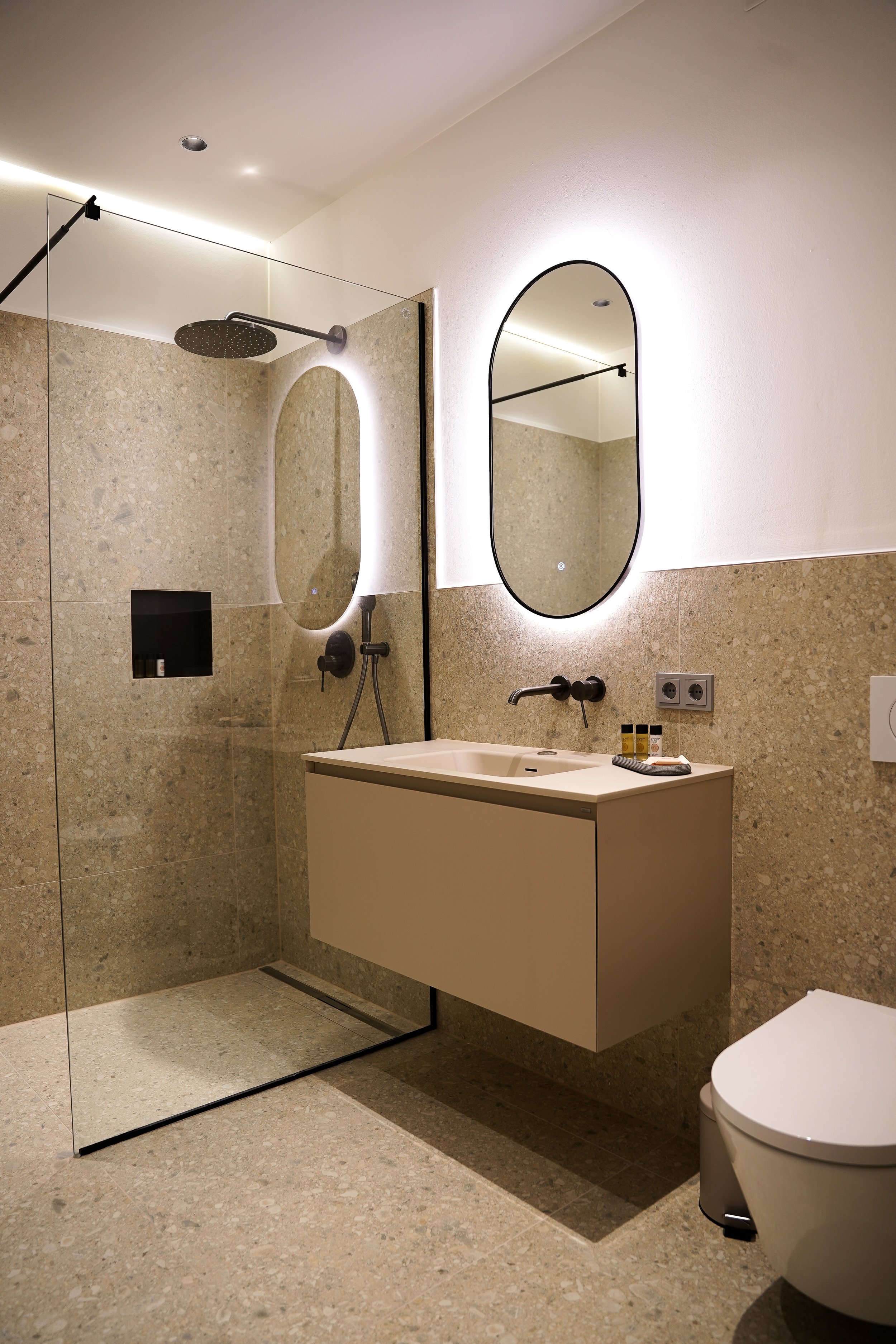 Moderner Badezimmer mit begehbarer Dusche, ovalem Spiegel mit Beleuchtung, schwebendem Waschtisch, schwarzen Armaturen und Wandsteckdosen.