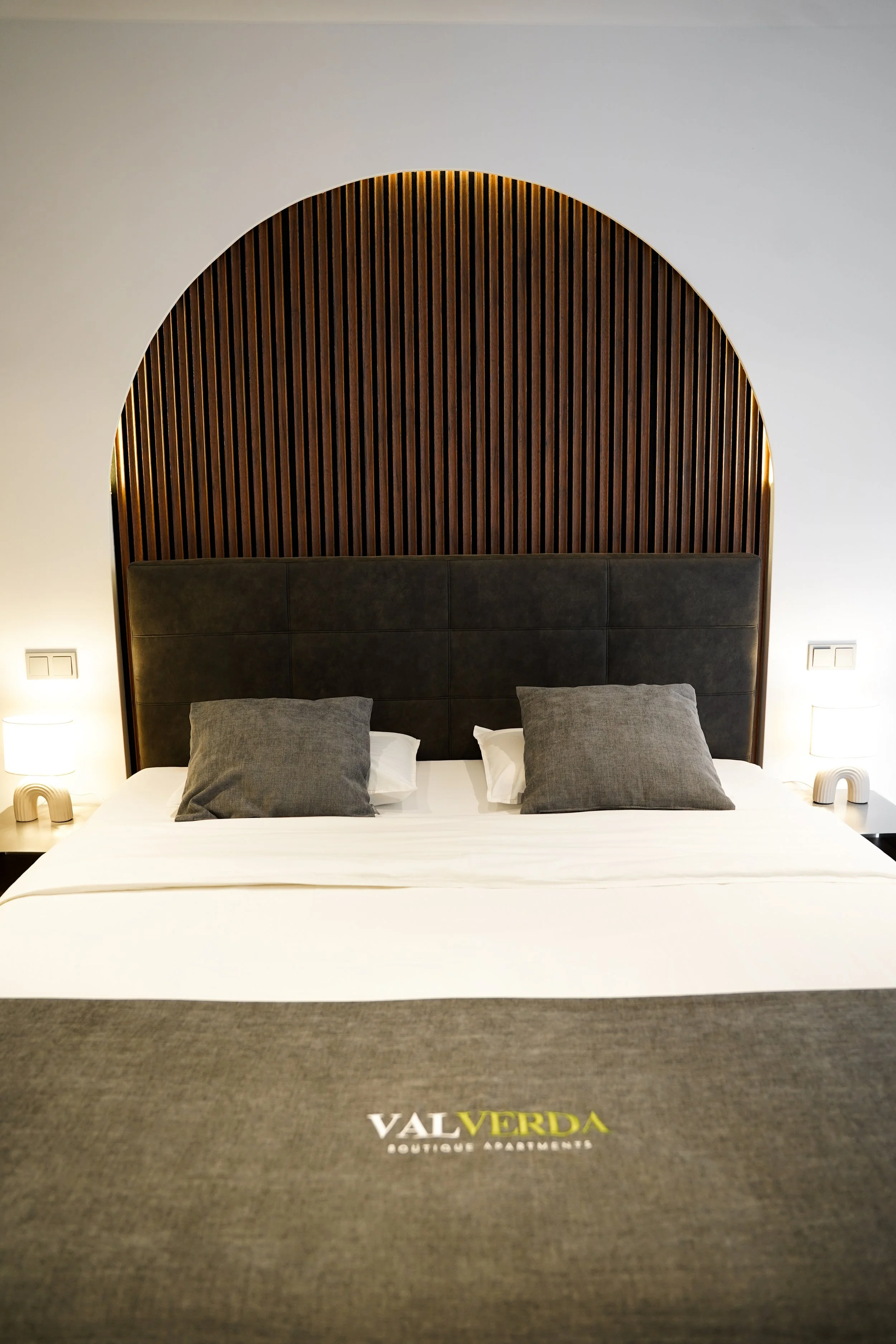 Modernes Hotelzimmerbett mit schwarzen Kopfteil, grauen Kissen, weißen Bettlaken und einer grauen Tagesdecke mit dem logo 'VAL VERDA'.