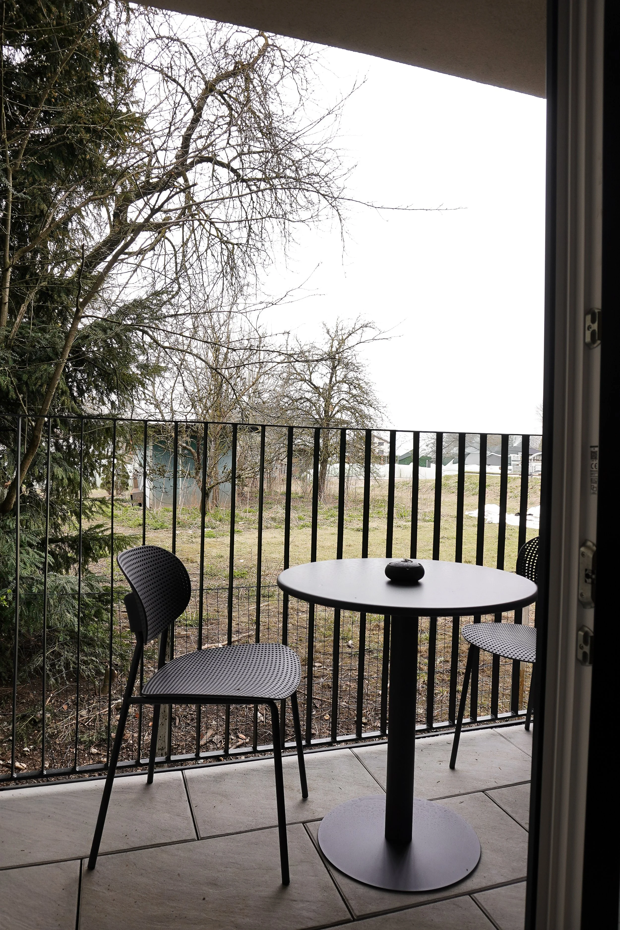 Balkon mit zwei schwarzen Stühlen, einem runden Tisch und einer Steinchen in der Mitte, Blick auf einen Garten mit Bäumen und einer Hütte bei bewölktem Himmel.